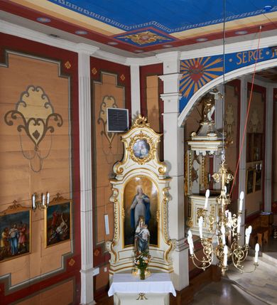 Zdjęcie nr 38: Kościół usytuowany jest w centrum wsi na niewielkim wzniesieniu, orientowany, drewniany, o konstrukcji zrębowej, oszalowany. Jego bryłę tworzy trójbocznie zamknięte prezbiterium, jednonawowy korpus, zakrystia skomunikowana ze skarbcem oraz kruchty od południa i zachodu. Całość pokrywają oddzielne dla nawy i prezbiterium dachy, nad nawą znajduje się wieżyczka na sygnaturkę. Wnętrza niższego, trójbocznie zamkniętego prezbiterium i szerszej, prawie kwadratowej nawy nakryte są stropem. Ściany kościoła zostały podzielone na przęsła kanelowanymi pilastrami ustawionymi na cokołach i zwieńczone profilowanym gzymsem, wyłamującym się na kapitelach. W prezbiterium znajdują się dwa okna od południa i dwa w bocznych ścianach wschodniego zamknięcia; korpus jest oświetlony od południa dwoma oknami. Wszystkie okna zamknięte są łukiem odcinkowym i umieszczone w prostokątnych obramieniach. Przestrzeń między prezbiterium a nawą jest nieznacznie zawężona wydatnymi pilastrami ustawionymi pod arkadą tęczową o łuku koszowym. W środkowym przęśle prezbiterium znajduje się wejście do północnej zakrystii, z profilowanym portalem zamkniętym łukiem trójlistnie ściętym. Na osi ściany południowej umieszczono prostokątny portal, ozdobiony od strony kruchty profilowaniem, a w nadprożu zdwojonym łukiem w  „ośli grzbiet”. Pod nim ornament arkadkowy i wyryta data „1547”. Główne wejście w ścianie zachodniej obejmuje profilowany od strony kruchty portal, który podobnie jak poprzedni zamknięty jest zdwojonym łukiem w „ośli grzbiet”. Chór muzyczny opiera się na dwóch kwadratowych słupach ustawionych diagonalnie (górą dekoracyjnie wycinanych), z parapetem o linii wklęsło-wypukłej. W południowo-zachodnim narożu znajdują się schody na chór osłonięte deskami. Zakrystia jest założona na rzucie prostokąta, przekryta pułapem, z lisicami na ścianie południowej i zachodniej. W osi ściany wschodniej zakrystii umieszczono okno zamknięte łukiem odcinkowym, a w narożu południowo-zachodnim schody na ambonę. W zachodniej ścianie zakrystii znajduje się prostokątne przejście do wąskiego przedsionka i dalej do dawnego skarbca przylegającego do korpusu. Kruchty są założone na rzucie prostokąta, wzdłużnie przylegającego do korpusu i przykryte stropem. Naprzeciw fasady ustawiono wolnostojącą, murowaną dzwonnicę.