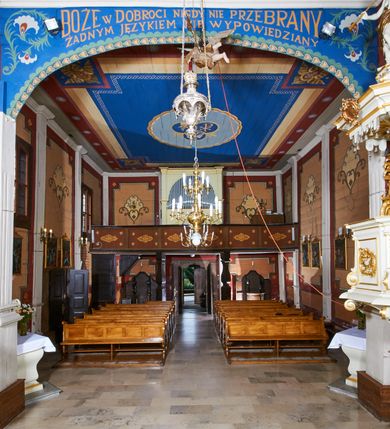 Zdjęcie nr 55: Kościół usytuowany jest w centrum wsi na niewielkim wzniesieniu, orientowany, drewniany, o konstrukcji zrębowej, oszalowany. Jego bryłę tworzy trójbocznie zamknięte prezbiterium, jednonawowy korpus, zakrystia skomunikowana ze skarbcem oraz kruchty od południa i zachodu. Całość pokrywają oddzielne dla nawy i prezbiterium dachy, nad nawą znajduje się wieżyczka na sygnaturkę. Wnętrza niższego, trójbocznie zamkniętego prezbiterium i szerszej, prawie kwadratowej nawy nakryte są stropem. Ściany kościoła zostały podzielone na przęsła kanelowanymi pilastrami ustawionymi na cokołach i zwieńczone profilowanym gzymsem, wyłamującym się na kapitelach. W prezbiterium znajdują się dwa okna od południa i dwa w bocznych ścianach wschodniego zamknięcia; korpus jest oświetlony od południa dwoma oknami. Wszystkie okna zamknięte są łukiem odcinkowym i umieszczone w prostokątnych obramieniach. Przestrzeń między prezbiterium a nawą jest nieznacznie zawężona wydatnymi pilastrami ustawionymi pod arkadą tęczową o łuku koszowym. W środkowym przęśle prezbiterium znajduje się wejście do północnej zakrystii, z profilowanym portalem zamkniętym łukiem trójlistnie ściętym. Na osi ściany południowej umieszczono prostokątny portal, ozdobiony od strony kruchty profilowaniem, a w nadprożu zdwojonym łukiem w  „ośli grzbiet”. Pod nim ornament arkadkowy i wyryta data „1547”. Główne wejście w ścianie zachodniej obejmuje profilowany od strony kruchty portal, który podobnie jak poprzedni zamknięty jest zdwojonym łukiem w „ośli grzbiet”. Chór muzyczny opiera się na dwóch kwadratowych słupach ustawionych diagonalnie (górą dekoracyjnie wycinanych), z parapetem o linii wklęsło-wypukłej. W południowo-zachodnim narożu znajdują się schody na chór osłonięte deskami. Zakrystia jest założona na rzucie prostokąta, przekryta pułapem, z lisicami na ścianie południowej i zachodniej. W osi ściany wschodniej zakrystii umieszczono okno zamknięte łukiem odcinkowym, a w narożu południowo-zachodnim schody na ambonę. W zachodniej ścianie zakrystii znajduje się prostokątne przejście do wąskiego przedsionka i dalej do dawnego skarbca przylegającego do korpusu. Kruchty są założone na rzucie prostokąta, wzdłużnie przylegającego do korpusu i przykryte stropem. Naprzeciw fasady ustawiono wolnostojącą, murowaną dzwonnicę.