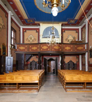 Zdjęcie nr 58: Kościół usytuowany jest w centrum wsi na niewielkim wzniesieniu, orientowany, drewniany, o konstrukcji zrębowej, oszalowany. Jego bryłę tworzy trójbocznie zamknięte prezbiterium, jednonawowy korpus, zakrystia skomunikowana ze skarbcem oraz kruchty od południa i zachodu. Całość pokrywają oddzielne dla nawy i prezbiterium dachy, nad nawą znajduje się wieżyczka na sygnaturkę. Wnętrza niższego, trójbocznie zamkniętego prezbiterium i szerszej, prawie kwadratowej nawy nakryte są stropem. Ściany kościoła zostały podzielone na przęsła kanelowanymi pilastrami ustawionymi na cokołach i zwieńczone profilowanym gzymsem, wyłamującym się na kapitelach. W prezbiterium znajdują się dwa okna od południa i dwa w bocznych ścianach wschodniego zamknięcia; korpus jest oświetlony od południa dwoma oknami. Wszystkie okna zamknięte są łukiem odcinkowym i umieszczone w prostokątnych obramieniach. Przestrzeń między prezbiterium a nawą jest nieznacznie zawężona wydatnymi pilastrami ustawionymi pod arkadą tęczową o łuku koszowym. W środkowym przęśle prezbiterium znajduje się wejście do północnej zakrystii, z profilowanym portalem zamkniętym łukiem trójlistnie ściętym. Na osi ściany południowej umieszczono prostokątny portal, ozdobiony od strony kruchty profilowaniem, a w nadprożu zdwojonym łukiem w  „ośli grzbiet”. Pod nim ornament arkadkowy i wyryta data „1547”. Główne wejście w ścianie zachodniej obejmuje profilowany od strony kruchty portal, który podobnie jak poprzedni zamknięty jest zdwojonym łukiem w „ośli grzbiet”. Chór muzyczny opiera się na dwóch kwadratowych słupach ustawionych diagonalnie (górą dekoracyjnie wycinanych), z parapetem o linii wklęsło-wypukłej. W południowo-zachodnim narożu znajdują się schody na chór osłonięte deskami. Zakrystia jest założona na rzucie prostokąta, przekryta pułapem, z lisicami na ścianie południowej i zachodniej. W osi ściany wschodniej zakrystii umieszczono okno zamknięte łukiem odcinkowym, a w narożu południowo-zachodnim schody na ambonę. W zachodniej ścianie zakrystii znajduje się prostokątne przejście do wąskiego przedsionka i dalej do dawnego skarbca przylegającego do korpusu. Kruchty są założone na rzucie prostokąta, wzdłużnie przylegającego do korpusu i przykryte stropem. Naprzeciw fasady ustawiono wolnostojącą, murowaną dzwonnicę.