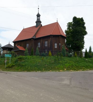 Zdjęcie nr 7: Kościół usytuowany jest w centrum wsi na niewielkim wzniesieniu, orientowany, drewniany, o konstrukcji zrębowej, oszalowany. Jego bryłę tworzy trójbocznie zamknięte prezbiterium, jednonawowy korpus, zakrystia skomunikowana ze skarbcem oraz kruchty od południa i zachodu. Całość pokrywają oddzielne dla nawy i prezbiterium dachy, nad nawą znajduje się wieżyczka na sygnaturkę. Wnętrza niższego, trójbocznie zamkniętego prezbiterium i szerszej, prawie kwadratowej nawy nakryte są stropem. Ściany kościoła zostały podzielone na przęsła kanelowanymi pilastrami ustawionymi na cokołach i zwieńczone profilowanym gzymsem, wyłamującym się na kapitelach. W prezbiterium znajdują się dwa okna od południa i dwa w bocznych ścianach wschodniego zamknięcia; korpus jest oświetlony od południa dwoma oknami. Wszystkie okna zamknięte są łukiem odcinkowym i umieszczone w prostokątnych obramieniach. Przestrzeń między prezbiterium a nawą jest nieznacznie zawężona wydatnymi pilastrami ustawionymi pod arkadą tęczową o łuku koszowym. W środkowym przęśle prezbiterium znajduje się wejście do północnej zakrystii, z profilowanym portalem zamkniętym łukiem trójlistnie ściętym. Na osi ściany południowej umieszczono prostokątny portal, ozdobiony od strony kruchty profilowaniem, a w nadprożu zdwojonym łukiem w  „ośli grzbiet”. Pod nim ornament arkadkowy i wyryta data „1547”. Główne wejście w ścianie zachodniej obejmuje profilowany od strony kruchty portal, który podobnie jak poprzedni zamknięty jest zdwojonym łukiem w „ośli grzbiet”. Chór muzyczny opiera się na dwóch kwadratowych słupach ustawionych diagonalnie (górą dekoracyjnie wycinanych), z parapetem o linii wklęsło-wypukłej. W południowo-zachodnim narożu znajdują się schody na chór osłonięte deskami. Zakrystia jest założona na rzucie prostokąta, przekryta pułapem, z lisicami na ścianie południowej i zachodniej. W osi ściany wschodniej zakrystii umieszczono okno zamknięte łukiem odcinkowym, a w narożu południowo-zachodnim schody na ambonę. W zachodniej ścianie zakrystii znajduje się prostokątne przejście do wąskiego przedsionka i dalej do dawnego skarbca przylegającego do korpusu. Kruchty są założone na rzucie prostokąta, wzdłużnie przylegającego do korpusu i przykryte stropem. Naprzeciw fasady ustawiono wolnostojącą, murowaną dzwonnicę.