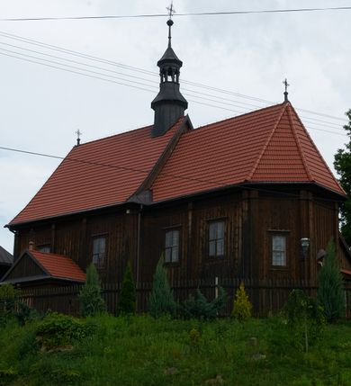 Zdjęcie nr 12: Kościół usytuowany jest w centrum wsi na niewielkim wzniesieniu, orientowany, drewniany, o konstrukcji zrębowej, oszalowany. Jego bryłę tworzy trójbocznie zamknięte prezbiterium, jednonawowy korpus, zakrystia skomunikowana ze skarbcem oraz kruchty od południa i zachodu. Całość pokrywają oddzielne dla nawy i prezbiterium dachy, nad nawą znajduje się wieżyczka na sygnaturkę. Wnętrza niższego, trójbocznie zamkniętego prezbiterium i szerszej, prawie kwadratowej nawy nakryte są stropem. Ściany kościoła zostały podzielone na przęsła kanelowanymi pilastrami ustawionymi na cokołach i zwieńczone profilowanym gzymsem, wyłamującym się na kapitelach. W prezbiterium znajdują się dwa okna od południa i dwa w bocznych ścianach wschodniego zamknięcia; korpus jest oświetlony od południa dwoma oknami. Wszystkie okna zamknięte są łukiem odcinkowym i umieszczone w prostokątnych obramieniach. Przestrzeń między prezbiterium a nawą jest nieznacznie zawężona wydatnymi pilastrami ustawionymi pod arkadą tęczową o łuku koszowym. W środkowym przęśle prezbiterium znajduje się wejście do północnej zakrystii, z profilowanym portalem zamkniętym łukiem trójlistnie ściętym. Na osi ściany południowej umieszczono prostokątny portal, ozdobiony od strony kruchty profilowaniem, a w nadprożu zdwojonym łukiem w  „ośli grzbiet”. Pod nim ornament arkadkowy i wyryta data „1547”. Główne wejście w ścianie zachodniej obejmuje profilowany od strony kruchty portal, który podobnie jak poprzedni zamknięty jest zdwojonym łukiem w „ośli grzbiet”. Chór muzyczny opiera się na dwóch kwadratowych słupach ustawionych diagonalnie (górą dekoracyjnie wycinanych), z parapetem o linii wklęsło-wypukłej. W południowo-zachodnim narożu znajdują się schody na chór osłonięte deskami. Zakrystia jest założona na rzucie prostokąta, przekryta pułapem, z lisicami na ścianie południowej i zachodniej. W osi ściany wschodniej zakrystii umieszczono okno zamknięte łukiem odcinkowym, a w narożu południowo-zachodnim schody na ambonę. W zachodniej ścianie zakrystii znajduje się prostokątne przejście do wąskiego przedsionka i dalej do dawnego skarbca przylegającego do korpusu. Kruchty są założone na rzucie prostokąta, wzdłużnie przylegającego do korpusu i przykryte stropem. Naprzeciw fasady ustawiono wolnostojącą, murowaną dzwonnicę.