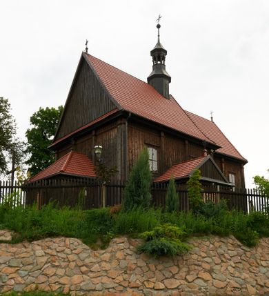 Zdjęcie nr 1: Kościół usytuowany jest w centrum wsi na niewielkim wzniesieniu, orientowany, drewniany, o konstrukcji zrębowej, oszalowany. Jego bryłę tworzy trójbocznie zamknięte prezbiterium, jednonawowy korpus, zakrystia skomunikowana ze skarbcem oraz kruchty od południa i zachodu. Całość pokrywają oddzielne dla nawy i prezbiterium dachy, nad nawą znajduje się wieżyczka na sygnaturkę. Wnętrza niższego, trójbocznie zamkniętego prezbiterium i szerszej, prawie kwadratowej nawy nakryte są stropem. Ściany kościoła zostały podzielone na przęsła kanelowanymi pilastrami ustawionymi na cokołach i zwieńczone profilowanym gzymsem, wyłamującym się na kapitelach. W prezbiterium znajdują się dwa okna od południa i dwa w bocznych ścianach wschodniego zamknięcia; korpus jest oświetlony od południa dwoma oknami. Wszystkie okna zamknięte są łukiem odcinkowym i umieszczone w prostokątnych obramieniach. Przestrzeń między prezbiterium a nawą jest nieznacznie zawężona wydatnymi pilastrami ustawionymi pod arkadą tęczową o łuku koszowym. W środkowym przęśle prezbiterium znajduje się wejście do północnej zakrystii, z profilowanym portalem zamkniętym łukiem trójlistnie ściętym. Na osi ściany południowej umieszczono prostokątny portal, ozdobiony od strony kruchty profilowaniem, a w nadprożu zdwojonym łukiem w  „ośli grzbiet”. Pod nim ornament arkadkowy i wyryta data „1547”. Główne wejście w ścianie zachodniej obejmuje profilowany od strony kruchty portal, który podobnie jak poprzedni zamknięty jest zdwojonym łukiem w „ośli grzbiet”. Chór muzyczny opiera się na dwóch kwadratowych słupach ustawionych diagonalnie (górą dekoracyjnie wycinanych), z parapetem o linii wklęsło-wypukłej. W południowo-zachodnim narożu znajdują się schody na chór osłonięte deskami. Zakrystia jest założona na rzucie prostokąta, przekryta pułapem, z lisicami na ścianie południowej i zachodniej. W osi ściany wschodniej zakrystii umieszczono okno zamknięte łukiem odcinkowym, a w narożu południowo-zachodnim schody na ambonę. W zachodniej ścianie zakrystii znajduje się prostokątne przejście do wąskiego przedsionka i dalej do dawnego skarbca przylegającego do korpusu. Kruchty są założone na rzucie prostokąta, wzdłużnie przylegającego do korpusu i przykryte stropem. Naprzeciw fasady ustawiono wolnostojącą, murowaną dzwonnicę.