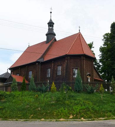 Zdjęcie nr 9: Kościół usytuowany jest w centrum wsi na niewielkim wzniesieniu, orientowany, drewniany, o konstrukcji zrębowej, oszalowany. Jego bryłę tworzy trójbocznie zamknięte prezbiterium, jednonawowy korpus, zakrystia skomunikowana ze skarbcem oraz kruchty od południa i zachodu. Całość pokrywają oddzielne dla nawy i prezbiterium dachy, nad nawą znajduje się wieżyczka na sygnaturkę. Wnętrza niższego, trójbocznie zamkniętego prezbiterium i szerszej, prawie kwadratowej nawy nakryte są stropem. Ściany kościoła zostały podzielone na przęsła kanelowanymi pilastrami ustawionymi na cokołach i zwieńczone profilowanym gzymsem, wyłamującym się na kapitelach. W prezbiterium znajdują się dwa okna od południa i dwa w bocznych ścianach wschodniego zamknięcia; korpus jest oświetlony od południa dwoma oknami. Wszystkie okna zamknięte są łukiem odcinkowym i umieszczone w prostokątnych obramieniach. Przestrzeń między prezbiterium a nawą jest nieznacznie zawężona wydatnymi pilastrami ustawionymi pod arkadą tęczową o łuku koszowym. W środkowym przęśle prezbiterium znajduje się wejście do północnej zakrystii, z profilowanym portalem zamkniętym łukiem trójlistnie ściętym. Na osi ściany południowej umieszczono prostokątny portal, ozdobiony od strony kruchty profilowaniem, a w nadprożu zdwojonym łukiem w  „ośli grzbiet”. Pod nim ornament arkadkowy i wyryta data „1547”. Główne wejście w ścianie zachodniej obejmuje profilowany od strony kruchty portal, który podobnie jak poprzedni zamknięty jest zdwojonym łukiem w „ośli grzbiet”. Chór muzyczny opiera się na dwóch kwadratowych słupach ustawionych diagonalnie (górą dekoracyjnie wycinanych), z parapetem o linii wklęsło-wypukłej. W południowo-zachodnim narożu znajdują się schody na chór osłonięte deskami. Zakrystia jest założona na rzucie prostokąta, przekryta pułapem, z lisicami na ścianie południowej i zachodniej. W osi ściany wschodniej zakrystii umieszczono okno zamknięte łukiem odcinkowym, a w narożu południowo-zachodnim schody na ambonę. W zachodniej ścianie zakrystii znajduje się prostokątne przejście do wąskiego przedsionka i dalej do dawnego skarbca przylegającego do korpusu. Kruchty są założone na rzucie prostokąta, wzdłużnie przylegającego do korpusu i przykryte stropem. Naprzeciw fasady ustawiono wolnostojącą, murowaną dzwonnicę.