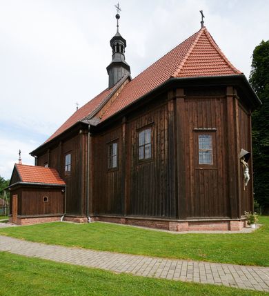 Zdjęcie nr 62: Kościół usytuowany jest w centrum wsi na niewielkim wzniesieniu, orientowany, drewniany, o konstrukcji zrębowej, oszalowany. Jego bryłę tworzy trójbocznie zamknięte prezbiterium, jednonawowy korpus, zakrystia skomunikowana ze skarbcem oraz kruchty od południa i zachodu. Całość pokrywają oddzielne dla nawy i prezbiterium dachy, nad nawą znajduje się wieżyczka na sygnaturkę. Wnętrza niższego, trójbocznie zamkniętego prezbiterium i szerszej, prawie kwadratowej nawy nakryte są stropem. Ściany kościoła zostały podzielone na przęsła kanelowanymi pilastrami ustawionymi na cokołach i zwieńczone profilowanym gzymsem, wyłamującym się na kapitelach. W prezbiterium znajdują się dwa okna od południa i dwa w bocznych ścianach wschodniego zamknięcia; korpus jest oświetlony od południa dwoma oknami. Wszystkie okna zamknięte są łukiem odcinkowym i umieszczone w prostokątnych obramieniach. Przestrzeń między prezbiterium a nawą jest nieznacznie zawężona wydatnymi pilastrami ustawionymi pod arkadą tęczową o łuku koszowym. W środkowym przęśle prezbiterium znajduje się wejście do północnej zakrystii, z profilowanym portalem zamkniętym łukiem trójlistnie ściętym. Na osi ściany południowej umieszczono prostokątny portal, ozdobiony od strony kruchty profilowaniem, a w nadprożu zdwojonym łukiem w  „ośli grzbiet”. Pod nim ornament arkadkowy i wyryta data „1547”. Główne wejście w ścianie zachodniej obejmuje profilowany od strony kruchty portal, który podobnie jak poprzedni zamknięty jest zdwojonym łukiem w „ośli grzbiet”. Chór muzyczny opiera się na dwóch kwadratowych słupach ustawionych diagonalnie (górą dekoracyjnie wycinanych), z parapetem o linii wklęsło-wypukłej. W południowo-zachodnim narożu znajdują się schody na chór osłonięte deskami. Zakrystia jest założona na rzucie prostokąta, przekryta pułapem, z lisicami na ścianie południowej i zachodniej. W osi ściany wschodniej zakrystii umieszczono okno zamknięte łukiem odcinkowym, a w narożu południowo-zachodnim schody na ambonę. W zachodniej ścianie zakrystii znajduje się prostokątne przejście do wąskiego przedsionka i dalej do dawnego skarbca przylegającego do korpusu. Kruchty są założone na rzucie prostokąta, wzdłużnie przylegającego do korpusu i przykryte stropem. Naprzeciw fasady ustawiono wolnostojącą, murowaną dzwonnicę.