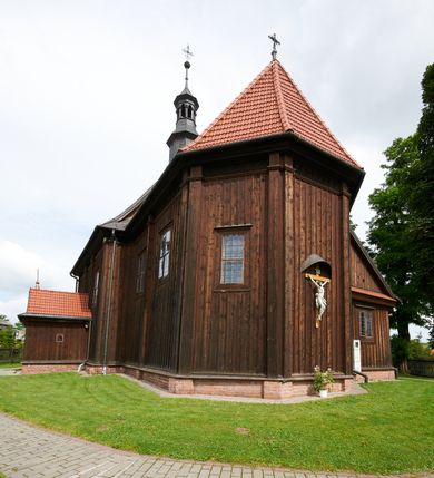 Zdjęcie nr 57: Kościół usytuowany jest w centrum wsi na niewielkim wzniesieniu, orientowany, drewniany, o konstrukcji zrębowej, oszalowany. Jego bryłę tworzy trójbocznie zamknięte prezbiterium, jednonawowy korpus, zakrystia skomunikowana ze skarbcem oraz kruchty od południa i zachodu. Całość pokrywają oddzielne dla nawy i prezbiterium dachy, nad nawą znajduje się wieżyczka na sygnaturkę. Wnętrza niższego, trójbocznie zamkniętego prezbiterium i szerszej, prawie kwadratowej nawy nakryte są stropem. Ściany kościoła zostały podzielone na przęsła kanelowanymi pilastrami ustawionymi na cokołach i zwieńczone profilowanym gzymsem, wyłamującym się na kapitelach. W prezbiterium znajdują się dwa okna od południa i dwa w bocznych ścianach wschodniego zamknięcia; korpus jest oświetlony od południa dwoma oknami. Wszystkie okna zamknięte są łukiem odcinkowym i umieszczone w prostokątnych obramieniach. Przestrzeń między prezbiterium a nawą jest nieznacznie zawężona wydatnymi pilastrami ustawionymi pod arkadą tęczową o łuku koszowym. W środkowym przęśle prezbiterium znajduje się wejście do północnej zakrystii, z profilowanym portalem zamkniętym łukiem trójlistnie ściętym. Na osi ściany południowej umieszczono prostokątny portal, ozdobiony od strony kruchty profilowaniem, a w nadprożu zdwojonym łukiem w  „ośli grzbiet”. Pod nim ornament arkadkowy i wyryta data „1547”. Główne wejście w ścianie zachodniej obejmuje profilowany od strony kruchty portal, który podobnie jak poprzedni zamknięty jest zdwojonym łukiem w „ośli grzbiet”. Chór muzyczny opiera się na dwóch kwadratowych słupach ustawionych diagonalnie (górą dekoracyjnie wycinanych), z parapetem o linii wklęsło-wypukłej. W południowo-zachodnim narożu znajdują się schody na chór osłonięte deskami. Zakrystia jest założona na rzucie prostokąta, przekryta pułapem, z lisicami na ścianie południowej i zachodniej. W osi ściany wschodniej zakrystii umieszczono okno zamknięte łukiem odcinkowym, a w narożu południowo-zachodnim schody na ambonę. W zachodniej ścianie zakrystii znajduje się prostokątne przejście do wąskiego przedsionka i dalej do dawnego skarbca przylegającego do korpusu. Kruchty są założone na rzucie prostokąta, wzdłużnie przylegającego do korpusu i przykryte stropem. Naprzeciw fasady ustawiono wolnostojącą, murowaną dzwonnicę.