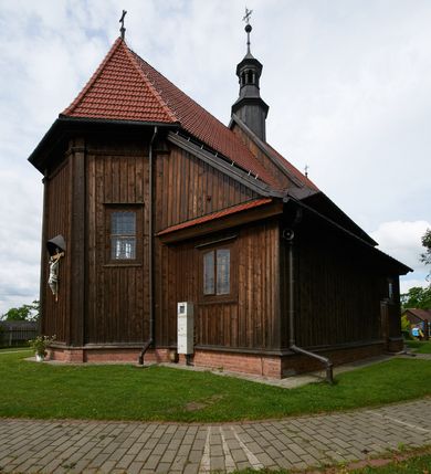 Zdjęcie nr 64: Kościół usytuowany jest w centrum wsi na niewielkim wzniesieniu, orientowany, drewniany, o konstrukcji zrębowej, oszalowany. Jego bryłę tworzy trójbocznie zamknięte prezbiterium, jednonawowy korpus, zakrystia skomunikowana ze skarbcem oraz kruchty od południa i zachodu. Całość pokrywają oddzielne dla nawy i prezbiterium dachy, nad nawą znajduje się wieżyczka na sygnaturkę. Wnętrza niższego, trójbocznie zamkniętego prezbiterium i szerszej, prawie kwadratowej nawy nakryte są stropem. Ściany kościoła zostały podzielone na przęsła kanelowanymi pilastrami ustawionymi na cokołach i zwieńczone profilowanym gzymsem, wyłamującym się na kapitelach. W prezbiterium znajdują się dwa okna od południa i dwa w bocznych ścianach wschodniego zamknięcia; korpus jest oświetlony od południa dwoma oknami. Wszystkie okna zamknięte są łukiem odcinkowym i umieszczone w prostokątnych obramieniach. Przestrzeń między prezbiterium a nawą jest nieznacznie zawężona wydatnymi pilastrami ustawionymi pod arkadą tęczową o łuku koszowym. W środkowym przęśle prezbiterium znajduje się wejście do północnej zakrystii, z profilowanym portalem zamkniętym łukiem trójlistnie ściętym. Na osi ściany południowej umieszczono prostokątny portal, ozdobiony od strony kruchty profilowaniem, a w nadprożu zdwojonym łukiem w  „ośli grzbiet”. Pod nim ornament arkadkowy i wyryta data „1547”. Główne wejście w ścianie zachodniej obejmuje profilowany od strony kruchty portal, który podobnie jak poprzedni zamknięty jest zdwojonym łukiem w „ośli grzbiet”. Chór muzyczny opiera się na dwóch kwadratowych słupach ustawionych diagonalnie (górą dekoracyjnie wycinanych), z parapetem o linii wklęsło-wypukłej. W południowo-zachodnim narożu znajdują się schody na chór osłonięte deskami. Zakrystia jest założona na rzucie prostokąta, przekryta pułapem, z lisicami na ścianie południowej i zachodniej. W osi ściany wschodniej zakrystii umieszczono okno zamknięte łukiem odcinkowym, a w narożu południowo-zachodnim schody na ambonę. W zachodniej ścianie zakrystii znajduje się prostokątne przejście do wąskiego przedsionka i dalej do dawnego skarbca przylegającego do korpusu. Kruchty są założone na rzucie prostokąta, wzdłużnie przylegającego do korpusu i przykryte stropem. Naprzeciw fasady ustawiono wolnostojącą, murowaną dzwonnicę.
