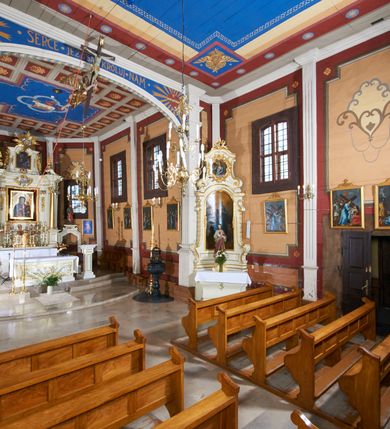 Zdjęcie nr 34: Kościół usytuowany jest w centrum wsi na niewielkim wzniesieniu, orientowany, drewniany, o konstrukcji zrębowej, oszalowany. Jego bryłę tworzy trójbocznie zamknięte prezbiterium, jednonawowy korpus, zakrystia skomunikowana ze skarbcem oraz kruchty od południa i zachodu. Całość pokrywają oddzielne dla nawy i prezbiterium dachy, nad nawą znajduje się wieżyczka na sygnaturkę. Wnętrza niższego, trójbocznie zamkniętego prezbiterium i szerszej, prawie kwadratowej nawy nakryte są stropem. Ściany kościoła zostały podzielone na przęsła kanelowanymi pilastrami ustawionymi na cokołach i zwieńczone profilowanym gzymsem, wyłamującym się na kapitelach. W prezbiterium znajdują się dwa okna od południa i dwa w bocznych ścianach wschodniego zamknięcia; korpus jest oświetlony od południa dwoma oknami. Wszystkie okna zamknięte są łukiem odcinkowym i umieszczone w prostokątnych obramieniach. Przestrzeń między prezbiterium a nawą jest nieznacznie zawężona wydatnymi pilastrami ustawionymi pod arkadą tęczową o łuku koszowym. W środkowym przęśle prezbiterium znajduje się wejście do północnej zakrystii, z profilowanym portalem zamkniętym łukiem trójlistnie ściętym. Na osi ściany południowej umieszczono prostokątny portal, ozdobiony od strony kruchty profilowaniem, a w nadprożu zdwojonym łukiem w  „ośli grzbiet”. Pod nim ornament arkadkowy i wyryta data „1547”. Główne wejście w ścianie zachodniej obejmuje profilowany od strony kruchty portal, który podobnie jak poprzedni zamknięty jest zdwojonym łukiem w „ośli grzbiet”. Chór muzyczny opiera się na dwóch kwadratowych słupach ustawionych diagonalnie (górą dekoracyjnie wycinanych), z parapetem o linii wklęsło-wypukłej. W południowo-zachodnim narożu znajdują się schody na chór osłonięte deskami. Zakrystia jest założona na rzucie prostokąta, przekryta pułapem, z lisicami na ścianie południowej i zachodniej. W osi ściany wschodniej zakrystii umieszczono okno zamknięte łukiem odcinkowym, a w narożu południowo-zachodnim schody na ambonę. W zachodniej ścianie zakrystii znajduje się prostokątne przejście do wąskiego przedsionka i dalej do dawnego skarbca przylegającego do korpusu. Kruchty są założone na rzucie prostokąta, wzdłużnie przylegającego do korpusu i przykryte stropem. Naprzeciw fasady ustawiono wolnostojącą, murowaną dzwonnicę.