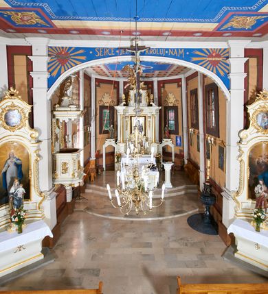 Zdjęcie nr 21: Kościół usytuowany jest w centrum wsi na niewielkim wzniesieniu, orientowany, drewniany, o konstrukcji zrębowej, oszalowany. Jego bryłę tworzy trójbocznie zamknięte prezbiterium, jednonawowy korpus, zakrystia skomunikowana ze skarbcem oraz kruchty od południa i zachodu. Całość pokrywają oddzielne dla nawy i prezbiterium dachy, nad nawą znajduje się wieżyczka na sygnaturkę. Wnętrza niższego, trójbocznie zamkniętego prezbiterium i szerszej, prawie kwadratowej nawy nakryte są stropem. Ściany kościoła zostały podzielone na przęsła kanelowanymi pilastrami ustawionymi na cokołach i zwieńczone profilowanym gzymsem, wyłamującym się na kapitelach. W prezbiterium znajdują się dwa okna od południa i dwa w bocznych ścianach wschodniego zamknięcia; korpus jest oświetlony od południa dwoma oknami. Wszystkie okna zamknięte są łukiem odcinkowym i umieszczone w prostokątnych obramieniach. Przestrzeń między prezbiterium a nawą jest nieznacznie zawężona wydatnymi pilastrami ustawionymi pod arkadą tęczową o łuku koszowym. W środkowym przęśle prezbiterium znajduje się wejście do północnej zakrystii, z profilowanym portalem zamkniętym łukiem trójlistnie ściętym. Na osi ściany południowej umieszczono prostokątny portal, ozdobiony od strony kruchty profilowaniem, a w nadprożu zdwojonym łukiem w  „ośli grzbiet”. Pod nim ornament arkadkowy i wyryta data „1547”. Główne wejście w ścianie zachodniej obejmuje profilowany od strony kruchty portal, który podobnie jak poprzedni zamknięty jest zdwojonym łukiem w „ośli grzbiet”. Chór muzyczny opiera się na dwóch kwadratowych słupach ustawionych diagonalnie (górą dekoracyjnie wycinanych), z parapetem o linii wklęsło-wypukłej. W południowo-zachodnim narożu znajdują się schody na chór osłonięte deskami. Zakrystia jest założona na rzucie prostokąta, przekryta pułapem, z lisicami na ścianie południowej i zachodniej. W osi ściany wschodniej zakrystii umieszczono okno zamknięte łukiem odcinkowym, a w narożu południowo-zachodnim schody na ambonę. W zachodniej ścianie zakrystii znajduje się prostokątne przejście do wąskiego przedsionka i dalej do dawnego skarbca przylegającego do korpusu. Kruchty są założone na rzucie prostokąta, wzdłużnie przylegającego do korpusu i przykryte stropem. Naprzeciw fasady ustawiono wolnostojącą, murowaną dzwonnicę.