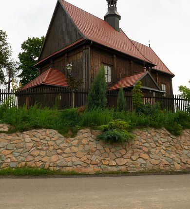 Zdjęcie nr 2: Kościół usytuowany jest w centrum wsi na niewielkim wzniesieniu, orientowany, drewniany, o konstrukcji zrębowej, oszalowany. Jego bryłę tworzy trójbocznie zamknięte prezbiterium, jednonawowy korpus, zakrystia skomunikowana ze skarbcem oraz kruchty od południa i zachodu. Całość pokrywają oddzielne dla nawy i prezbiterium dachy, nad nawą znajduje się wieżyczka na sygnaturkę. Wnętrza niższego, trójbocznie zamkniętego prezbiterium i szerszej, prawie kwadratowej nawy nakryte są stropem. Ściany kościoła zostały podzielone na przęsła kanelowanymi pilastrami ustawionymi na cokołach i zwieńczone profilowanym gzymsem, wyłamującym się na kapitelach. W prezbiterium znajdują się dwa okna od południa i dwa w bocznych ścianach wschodniego zamknięcia; korpus jest oświetlony od południa dwoma oknami. Wszystkie okna zamknięte są łukiem odcinkowym i umieszczone w prostokątnych obramieniach. Przestrzeń między prezbiterium a nawą jest nieznacznie zawężona wydatnymi pilastrami ustawionymi pod arkadą tęczową o łuku koszowym. W środkowym przęśle prezbiterium znajduje się wejście do północnej zakrystii, z profilowanym portalem zamkniętym łukiem trójlistnie ściętym. Na osi ściany południowej umieszczono prostokątny portal, ozdobiony od strony kruchty profilowaniem, a w nadprożu zdwojonym łukiem w  „ośli grzbiet”. Pod nim ornament arkadkowy i wyryta data „1547”. Główne wejście w ścianie zachodniej obejmuje profilowany od strony kruchty portal, który podobnie jak poprzedni zamknięty jest zdwojonym łukiem w „ośli grzbiet”. Chór muzyczny opiera się na dwóch kwadratowych słupach ustawionych diagonalnie (górą dekoracyjnie wycinanych), z parapetem o linii wklęsło-wypukłej. W południowo-zachodnim narożu znajdują się schody na chór osłonięte deskami. Zakrystia jest założona na rzucie prostokąta, przekryta pułapem, z lisicami na ścianie południowej i zachodniej. W osi ściany wschodniej zakrystii umieszczono okno zamknięte łukiem odcinkowym, a w narożu południowo-zachodnim schody na ambonę. W zachodniej ścianie zakrystii znajduje się prostokątne przejście do wąskiego przedsionka i dalej do dawnego skarbca przylegającego do korpusu. Kruchty są założone na rzucie prostokąta, wzdłużnie przylegającego do korpusu i przykryte stropem. Naprzeciw fasady ustawiono wolnostojącą, murowaną dzwonnicę.