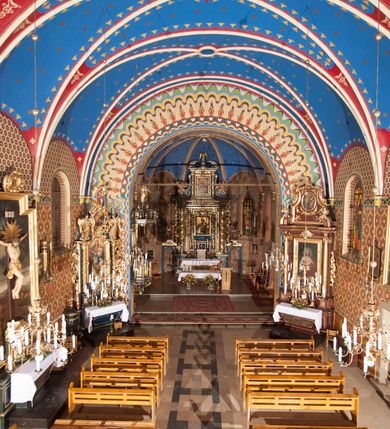 Zdjęcie nr 9: Kościół murowany, tynkowany, orientowany, jednonawowy. 
Prezbiterium prostokątne, zamknięte trójbocznie, od północy przylega zakrystia na planie prostokąta. Nawa szersza od prezbiterium, na rzucie prostokąta, od południa dostawiona kaplica Matki Boskiej na planie prostokąta, od zachodu wieża na planie kwadratu z dwoma aneksami po bokach. 
Prezbiterium dwuprzęsłowe, nawa trójprzęsłowa, nakryte sklepieniami krzyżowo-żebrowymi o gurtach spływających na profilowane wsporniki. Otwór tęczowy w formie półkolistej arkady. Parapet chóru muzycznego drewniany, wsparty na dwóch filarach.  Posadzka z płyt ceramicznych. 
Fasada wieżowa, jednoosiowa, trójkondygnacyjna, zwężająca się ku górze; dwie dolne kondygnacje na planie kwadratu, trzecia ośmioboku. Trzy ściany pierwszej kondygnacji wieży ujęte lizenami, zakończone profilowanym gzymsem z krenelażem oraz daszkiem pulpitowym. Na osi portal w formie stojącego prostokąta zamkniętego łukiem półkolistym, w opasce tynkowej z dwuskrzydłowymi drzwiami. Portal zamknięty od góry i po bokach uskokowym gzymsem z daszkiem pulpitowym. Nad portalem biforium. Trzy ściany drugiej kondygnacji wieży ujęte lizenami, zakończone profilowanym gzymsem z krenelażem oraz daszkiem pulpitowym. Na ścianach ślepe biforia we wnękach w formie stojącego prostokąta zamkniętego łukiem półkolistym. Trzecia kondygnacja zakończona profilowanym gzymsem z kostkowaniem, na ściankach otwory dzwonne w formie stojącego prostokąta zamkniętego łukiem półkolistym, naprzemiennie ślepe i otwarte. Wieża nakryta dachem ostrosłupowym. Po bokach wieży dwukondygnacyjne aneksy na planie wieloboku, przekryte daszkami wielospadowymi. 
Nawa i prezbiterium oszkarpowane w narożach jedno i dwuuskokowymi szkarpami nakrytymi daszkami. Na elewacji południowej pięć otworów okiennych w formie stojącego prostokąta zamkniętego łukiem półkolistym, z szerokim rozglifieniem. Na elewacji wschodniej dwa analogiczne okna, na północnej trzy. 
Nawa, kaplica i zakrystia nakryte dachami blaszanymi, dwuspadowymi, prezbiterium pięciospadowym. 