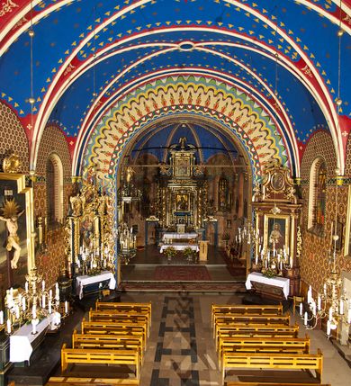 Zdjęcie nr 10: Kościół murowany, tynkowany, orientowany, jednonawowy. 
Prezbiterium prostokątne, zamknięte trójbocznie, od północy przylega zakrystia na planie prostokąta. Nawa szersza od prezbiterium, na rzucie prostokąta, od południa dostawiona kaplica Matki Boskiej na planie prostokąta, od zachodu wieża na planie kwadratu z dwoma aneksami po bokach. 
Prezbiterium dwuprzęsłowe, nawa trójprzęsłowa, nakryte sklepieniami krzyżowo-żebrowymi o gurtach spływających na profilowane wsporniki. Otwór tęczowy w formie półkolistej arkady. Parapet chóru muzycznego drewniany, wsparty na dwóch filarach.  Posadzka z płyt ceramicznych. 
Fasada wieżowa, jednoosiowa, trójkondygnacyjna, zwężająca się ku górze; dwie dolne kondygnacje na planie kwadratu, trzecia ośmioboku. Trzy ściany pierwszej kondygnacji wieży ujęte lizenami, zakończone profilowanym gzymsem z krenelażem oraz daszkiem pulpitowym. Na osi portal w formie stojącego prostokąta zamkniętego łukiem półkolistym, w opasce tynkowej z dwuskrzydłowymi drzwiami. Portal zamknięty od góry i po bokach uskokowym gzymsem z daszkiem pulpitowym. Nad portalem biforium. Trzy ściany drugiej kondygnacji wieży ujęte lizenami, zakończone profilowanym gzymsem z krenelażem oraz daszkiem pulpitowym. Na ścianach ślepe biforia we wnękach w formie stojącego prostokąta zamkniętego łukiem półkolistym. Trzecia kondygnacja zakończona profilowanym gzymsem z kostkowaniem, na ściankach otwory dzwonne w formie stojącego prostokąta zamkniętego łukiem półkolistym, naprzemiennie ślepe i otwarte. Wieża nakryta dachem ostrosłupowym. Po bokach wieży dwukondygnacyjne aneksy na planie wieloboku, przekryte daszkami wielospadowymi. 
Nawa i prezbiterium oszkarpowane w narożach jedno i dwuuskokowymi szkarpami nakrytymi daszkami. Na elewacji południowej pięć otworów okiennych w formie stojącego prostokąta zamkniętego łukiem półkolistym, z szerokim rozglifieniem. Na elewacji wschodniej dwa analogiczne okna, na północnej trzy. 
Nawa, kaplica i zakrystia nakryte dachami blaszanymi, dwuspadowymi, prezbiterium pięciospadowym. 