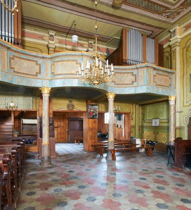 Zdjęcie nr 12: Kościół drewniany o konstrukcji zrębowej, na betonowym podmurowaniu przesłoniętym daszkiem gontowym, kruchta zachodnia o konstrukcji słupowo-ramowej. Elewacje jednolite, szalowane z listwowaniem, wzmocnione lisicami. Więźba dachowa drewniana, krokwiowa. Kościół orientowany, jednonawowy, nawa na rzucie prostokąta zbliżonego do kwadratu, prezbiterium nieco węższe, zakończone trójbocznie. Do wschodniej ściany prezbiterium przylega Ogrójec na rzucie prostokąta, od północy zakrystia na planie zbliżonym do trapezu. Do północnej ściany nawy przylega niewielka kruchta na rzucie prostokąta, od zachodu duża kruchta na planie prostokąta z pomieszczeniem nad nią, ze schodami drabiniastymi na chór, otwarta do nawy trzema dużymi otworami o wykroju prostokątnym. Wnętrze podzielone pilastrami podtrzymującymi architraw i fryz, wyłamane na ich osi. Pomiędzy nawą a prezbiterium prostokątny otwór tęczowy z prostą belką. Parapet chóru muzycznego w zachodniej części nawy, o wklęsło-wypukłym zarysie, podparty przez dwie kolumny i dwie półkolumny o wielobocznych trzonach. Otwory drzwiowe o prostokątnym wykroju zamknięte drzwiami spągowymi w północnej ścianie nawy oraz prezbiterium. Wnętrze nakryte pułapami deskowymi. Posadzka z płyt ceramicznych. 
Elewacja frontowa jednoosiowa, na osi wejście do kruchty z dwuskrzydłowymi drzwiami, w partii szczytu podwójne okno. W elewacji południowej cztery otwory okienne w formie stojącego prostokąta zamkniętego łukiem odcinkowym, dwa w nawie i dwa w prezbiterium oraz prostokątne, podwójne okno w kruchcie zachodniej, z krosnowymi oknami o drobnych podziałach kwaterowych. Elewacja północna z prostokątnym, podwójnym oknem w części kruchty zachodniej oraz z wejściami do kruchty północnej i do zakrystii o dwuskrzydłowych drzwiach. Na ścianie wschodniej prezbiterium Ogrójec w formie głębokiej szalowanej wnęki przesłoniętej daszkiem pulpitowym. Kościół nakryty dachami gontowymi dwuspadowymi, wyższym nad nawą, niższym nad prezbiterium, z kalenicą dekorowaną gontowym grzebieniem, nad zakrystią i kruchtą północną miękko modelowane dachy trójspadowe, kruchta zachodnia nakryta dachem dwuspadowym, półszczytowym. W połowie długości dachu nad nawą od strony prezbiterium wznosi się ponad kalenicą wieżyczka na sygnaturkę, na ośmiobocznym obitym blachą trzonie, z latarnią zwieńczoną niewielkim cebulastym hełmem.

 

