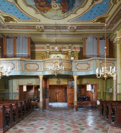 Zdjęcie nr 10: Kościół drewniany o konstrukcji zrębowej, na betonowym podmurowaniu przesłoniętym daszkiem gontowym, kruchta zachodnia o konstrukcji słupowo-ramowej. Elewacje jednolite, szalowane z listwowaniem, wzmocnione lisicami. Więźba dachowa drewniana, krokwiowa. Kościół orientowany, jednonawowy, nawa na rzucie prostokąta zbliżonego do kwadratu, prezbiterium nieco węższe, zakończone trójbocznie. Do wschodniej ściany prezbiterium przylega Ogrójec na rzucie prostokąta, od północy zakrystia na planie zbliżonym do trapezu. Do północnej ściany nawy przylega niewielka kruchta na rzucie prostokąta, od zachodu duża kruchta na planie prostokąta z pomieszczeniem nad nią, ze schodami drabiniastymi na chór, otwarta do nawy trzema dużymi otworami o wykroju prostokątnym. Wnętrze podzielone pilastrami podtrzymującymi architraw i fryz, wyłamane na ich osi. Pomiędzy nawą a prezbiterium prostokątny otwór tęczowy z prostą belką. Parapet chóru muzycznego w zachodniej części nawy, o wklęsło-wypukłym zarysie, podparty przez dwie kolumny i dwie półkolumny o wielobocznych trzonach. Otwory drzwiowe o prostokątnym wykroju zamknięte drzwiami spągowymi w północnej ścianie nawy oraz prezbiterium. Wnętrze nakryte pułapami deskowymi. Posadzka z płyt ceramicznych. 
Elewacja frontowa jednoosiowa, na osi wejście do kruchty z dwuskrzydłowymi drzwiami, w partii szczytu podwójne okno. W elewacji południowej cztery otwory okienne w formie stojącego prostokąta zamkniętego łukiem odcinkowym, dwa w nawie i dwa w prezbiterium oraz prostokątne, podwójne okno w kruchcie zachodniej, z krosnowymi oknami o drobnych podziałach kwaterowych. Elewacja północna z prostokątnym, podwójnym oknem w części kruchty zachodniej oraz z wejściami do kruchty północnej i do zakrystii o dwuskrzydłowych drzwiach. Na ścianie wschodniej prezbiterium Ogrójec w formie głębokiej szalowanej wnęki przesłoniętej daszkiem pulpitowym. Kościół nakryty dachami gontowymi dwuspadowymi, wyższym nad nawą, niższym nad prezbiterium, z kalenicą dekorowaną gontowym grzebieniem, nad zakrystią i kruchtą północną miękko modelowane dachy trójspadowe, kruchta zachodnia nakryta dachem dwuspadowym, półszczytowym. W połowie długości dachu nad nawą od strony prezbiterium wznosi się ponad kalenicą wieżyczka na sygnaturkę, na ośmiobocznym obitym blachą trzonie, z latarnią zwieńczoną niewielkim cebulastym hełmem.

 

