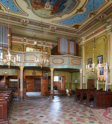 Zdjęcie nr 23: Kościół drewniany o konstrukcji zrębowej, na betonowym podmurowaniu przesłoniętym daszkiem gontowym, kruchta zachodnia o konstrukcji słupowo-ramowej. Elewacje jednolite, szalowane z listwowaniem, wzmocnione lisicami. Więźba dachowa drewniana, krokwiowa. Kościół orientowany, jednonawowy, nawa na rzucie prostokąta zbliżonego do kwadratu, prezbiterium nieco węższe, zakończone trójbocznie. Do wschodniej ściany prezbiterium przylega Ogrójec na rzucie prostokąta, od północy zakrystia na planie zbliżonym do trapezu. Do północnej ściany nawy przylega niewielka kruchta na rzucie prostokąta, od zachodu duża kruchta na planie prostokąta z pomieszczeniem nad nią, ze schodami drabiniastymi na chór, otwarta do nawy trzema dużymi otworami o wykroju prostokątnym. Wnętrze podzielone pilastrami podtrzymującymi architraw i fryz, wyłamane na ich osi. Pomiędzy nawą a prezbiterium prostokątny otwór tęczowy z prostą belką. Parapet chóru muzycznego w zachodniej części nawy, o wklęsło-wypukłym zarysie, podparty przez dwie kolumny i dwie półkolumny o wielobocznych trzonach. Otwory drzwiowe o prostokątnym wykroju zamknięte drzwiami spągowymi w północnej ścianie nawy oraz prezbiterium. Wnętrze nakryte pułapami deskowymi. Posadzka z płyt ceramicznych. 
Elewacja frontowa jednoosiowa, na osi wejście do kruchty z dwuskrzydłowymi drzwiami, w partii szczytu podwójne okno. W elewacji południowej cztery otwory okienne w formie stojącego prostokąta zamkniętego łukiem odcinkowym, dwa w nawie i dwa w prezbiterium oraz prostokątne, podwójne okno w kruchcie zachodniej, z krosnowymi oknami o drobnych podziałach kwaterowych. Elewacja północna z prostokątnym, podwójnym oknem w części kruchty zachodniej oraz z wejściami do kruchty północnej i do zakrystii o dwuskrzydłowych drzwiach. Na ścianie wschodniej prezbiterium Ogrójec w formie głębokiej szalowanej wnęki przesłoniętej daszkiem pulpitowym. Kościół nakryty dachami gontowymi dwuspadowymi, wyższym nad nawą, niższym nad prezbiterium, z kalenicą dekorowaną gontowym grzebieniem, nad zakrystią i kruchtą północną miękko modelowane dachy trójspadowe, kruchta zachodnia nakryta dachem dwuspadowym, półszczytowym. W połowie długości dachu nad nawą od strony prezbiterium wznosi się ponad kalenicą wieżyczka na sygnaturkę, na ośmiobocznym obitym blachą trzonie, z latarnią zwieńczoną niewielkim cebulastym hełmem.

 

