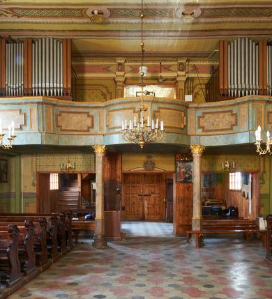 Zdjęcie nr 25: Kościół drewniany o konstrukcji zrębowej, na betonowym podmurowaniu przesłoniętym daszkiem gontowym, kruchta zachodnia o konstrukcji słupowo-ramowej. Elewacje jednolite, szalowane z listwowaniem, wzmocnione lisicami. Więźba dachowa drewniana, krokwiowa. Kościół orientowany, jednonawowy, nawa na rzucie prostokąta zbliżonego do kwadratu, prezbiterium nieco węższe, zakończone trójbocznie. Do wschodniej ściany prezbiterium przylega Ogrójec na rzucie prostokąta, od północy zakrystia na planie zbliżonym do trapezu. Do północnej ściany nawy przylega niewielka kruchta na rzucie prostokąta, od zachodu duża kruchta na planie prostokąta z pomieszczeniem nad nią, ze schodami drabiniastymi na chór, otwarta do nawy trzema dużymi otworami o wykroju prostokątnym. Wnętrze podzielone pilastrami podtrzymującymi architraw i fryz, wyłamane na ich osi. Pomiędzy nawą a prezbiterium prostokątny otwór tęczowy z prostą belką. Parapet chóru muzycznego w zachodniej części nawy, o wklęsło-wypukłym zarysie, podparty przez dwie kolumny i dwie półkolumny o wielobocznych trzonach. Otwory drzwiowe o prostokątnym wykroju zamknięte drzwiami spągowymi w północnej ścianie nawy oraz prezbiterium. Wnętrze nakryte pułapami deskowymi. Posadzka z płyt ceramicznych. 
Elewacja frontowa jednoosiowa, na osi wejście do kruchty z dwuskrzydłowymi drzwiami, w partii szczytu podwójne okno. W elewacji południowej cztery otwory okienne w formie stojącego prostokąta zamkniętego łukiem odcinkowym, dwa w nawie i dwa w prezbiterium oraz prostokątne, podwójne okno w kruchcie zachodniej, z krosnowymi oknami o drobnych podziałach kwaterowych. Elewacja północna z prostokątnym, podwójnym oknem w części kruchty zachodniej oraz z wejściami do kruchty północnej i do zakrystii o dwuskrzydłowych drzwiach. Na ścianie wschodniej prezbiterium Ogrójec w formie głębokiej szalowanej wnęki przesłoniętej daszkiem pulpitowym. Kościół nakryty dachami gontowymi dwuspadowymi, wyższym nad nawą, niższym nad prezbiterium, z kalenicą dekorowaną gontowym grzebieniem, nad zakrystią i kruchtą północną miękko modelowane dachy trójspadowe, kruchta zachodnia nakryta dachem dwuspadowym, półszczytowym. W połowie długości dachu nad nawą od strony prezbiterium wznosi się ponad kalenicą wieżyczka na sygnaturkę, na ośmiobocznym obitym blachą trzonie, z latarnią zwieńczoną niewielkim cebulastym hełmem.

 

