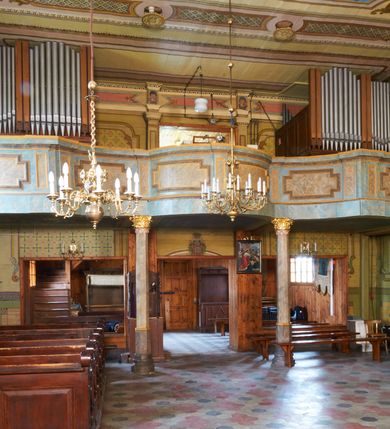 Zdjęcie nr 24: Kościół drewniany o konstrukcji zrębowej, na betonowym podmurowaniu przesłoniętym daszkiem gontowym, kruchta zachodnia o konstrukcji słupowo-ramowej. Elewacje jednolite, szalowane z listwowaniem, wzmocnione lisicami. Więźba dachowa drewniana, krokwiowa. Kościół orientowany, jednonawowy, nawa na rzucie prostokąta zbliżonego do kwadratu, prezbiterium nieco węższe, zakończone trójbocznie. Do wschodniej ściany prezbiterium przylega Ogrójec na rzucie prostokąta, od północy zakrystia na planie zbliżonym do trapezu. Do północnej ściany nawy przylega niewielka kruchta na rzucie prostokąta, od zachodu duża kruchta na planie prostokąta z pomieszczeniem nad nią, ze schodami drabiniastymi na chór, otwarta do nawy trzema dużymi otworami o wykroju prostokątnym. Wnętrze podzielone pilastrami podtrzymującymi architraw i fryz, wyłamane na ich osi. Pomiędzy nawą a prezbiterium prostokątny otwór tęczowy z prostą belką. Parapet chóru muzycznego w zachodniej części nawy, o wklęsło-wypukłym zarysie, podparty przez dwie kolumny i dwie półkolumny o wielobocznych trzonach. Otwory drzwiowe o prostokątnym wykroju zamknięte drzwiami spągowymi w północnej ścianie nawy oraz prezbiterium. Wnętrze nakryte pułapami deskowymi. Posadzka z płyt ceramicznych. 
Elewacja frontowa jednoosiowa, na osi wejście do kruchty z dwuskrzydłowymi drzwiami, w partii szczytu podwójne okno. W elewacji południowej cztery otwory okienne w formie stojącego prostokąta zamkniętego łukiem odcinkowym, dwa w nawie i dwa w prezbiterium oraz prostokątne, podwójne okno w kruchcie zachodniej, z krosnowymi oknami o drobnych podziałach kwaterowych. Elewacja północna z prostokątnym, podwójnym oknem w części kruchty zachodniej oraz z wejściami do kruchty północnej i do zakrystii o dwuskrzydłowych drzwiach. Na ścianie wschodniej prezbiterium Ogrójec w formie głębokiej szalowanej wnęki przesłoniętej daszkiem pulpitowym. Kościół nakryty dachami gontowymi dwuspadowymi, wyższym nad nawą, niższym nad prezbiterium, z kalenicą dekorowaną gontowym grzebieniem, nad zakrystią i kruchtą północną miękko modelowane dachy trójspadowe, kruchta zachodnia nakryta dachem dwuspadowym, półszczytowym. W połowie długości dachu nad nawą od strony prezbiterium wznosi się ponad kalenicą wieżyczka na sygnaturkę, na ośmiobocznym obitym blachą trzonie, z latarnią zwieńczoną niewielkim cebulastym hełmem.

 

