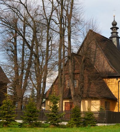 Zdjęcie nr 3: Kościół drewniany o konstrukcji zrębowej, na betonowym podmurowaniu przesłoniętym daszkiem gontowym, kruchta zachodnia o konstrukcji słupowo-ramowej. Elewacje jednolite, szalowane z listwowaniem, wzmocnione lisicami. Więźba dachowa drewniana, krokwiowa. Kościół orientowany, jednonawowy, nawa na rzucie prostokąta zbliżonego do kwadratu, prezbiterium nieco węższe, zakończone trójbocznie. Do wschodniej ściany prezbiterium przylega Ogrójec na rzucie prostokąta, od północy zakrystia na planie zbliżonym do trapezu. Do północnej ściany nawy przylega niewielka kruchta na rzucie prostokąta, od zachodu duża kruchta na planie prostokąta z pomieszczeniem nad nią, ze schodami drabiniastymi na chór, otwarta do nawy trzema dużymi otworami o wykroju prostokątnym. Wnętrze podzielone pilastrami podtrzymującymi architraw i fryz, wyłamane na ich osi. Pomiędzy nawą a prezbiterium prostokątny otwór tęczowy z prostą belką. Parapet chóru muzycznego w zachodniej części nawy, o wklęsło-wypukłym zarysie, podparty przez dwie kolumny i dwie półkolumny o wielobocznych trzonach. Otwory drzwiowe o prostokątnym wykroju zamknięte drzwiami spągowymi w północnej ścianie nawy oraz prezbiterium. Wnętrze nakryte pułapami deskowymi. Posadzka z płyt ceramicznych. 
Elewacja frontowa jednoosiowa, na osi wejście do kruchty z dwuskrzydłowymi drzwiami, w partii szczytu podwójne okno. W elewacji południowej cztery otwory okienne w formie stojącego prostokąta zamkniętego łukiem odcinkowym, dwa w nawie i dwa w prezbiterium oraz prostokątne, podwójne okno w kruchcie zachodniej, z krosnowymi oknami o drobnych podziałach kwaterowych. Elewacja północna z prostokątnym, podwójnym oknem w części kruchty zachodniej oraz z wejściami do kruchty północnej i do zakrystii o dwuskrzydłowych drzwiach. Na ścianie wschodniej prezbiterium Ogrójec w formie głębokiej szalowanej wnęki przesłoniętej daszkiem pulpitowym. Kościół nakryty dachami gontowymi dwuspadowymi, wyższym nad nawą, niższym nad prezbiterium, z kalenicą dekorowaną gontowym grzebieniem, nad zakrystią i kruchtą północną miękko modelowane dachy trójspadowe, kruchta zachodnia nakryta dachem dwuspadowym, półszczytowym. W połowie długości dachu nad nawą od strony prezbiterium wznosi się ponad kalenicą wieżyczka na sygnaturkę, na ośmiobocznym obitym blachą trzonie, z latarnią zwieńczoną niewielkim cebulastym hełmem.

 

