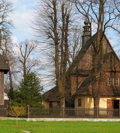 Zdjęcie nr 53: Kościół drewniany o konstrukcji zrębowej, na betonowym podmurowaniu przesłoniętym daszkiem gontowym, kruchta zachodnia o konstrukcji słupowo-ramowej. Elewacje jednolite, szalowane z listwowaniem, wzmocnione lisicami. Więźba dachowa drewniana, krokwiowa. Kościół orientowany, jednonawowy, nawa na rzucie prostokąta zbliżonego do kwadratu, prezbiterium nieco węższe, zakończone trójbocznie. Do wschodniej ściany prezbiterium przylega Ogrójec na rzucie prostokąta, od północy zakrystia na planie zbliżonym do trapezu. Do północnej ściany nawy przylega niewielka kruchta na rzucie prostokąta, od zachodu duża kruchta na planie prostokąta z pomieszczeniem nad nią, ze schodami drabiniastymi na chór, otwarta do nawy trzema dużymi otworami o wykroju prostokątnym. Wnętrze podzielone pilastrami podtrzymującymi architraw i fryz, wyłamane na ich osi. Pomiędzy nawą a prezbiterium prostokątny otwór tęczowy z prostą belką. Parapet chóru muzycznego w zachodniej części nawy, o wklęsło-wypukłym zarysie, podparty przez dwie kolumny i dwie półkolumny o wielobocznych trzonach. Otwory drzwiowe o prostokątnym wykroju zamknięte drzwiami spągowymi w północnej ścianie nawy oraz prezbiterium. Wnętrze nakryte pułapami deskowymi. Posadzka z płyt ceramicznych. 
Elewacja frontowa jednoosiowa, na osi wejście do kruchty z dwuskrzydłowymi drzwiami, w partii szczytu podwójne okno. W elewacji południowej cztery otwory okienne w formie stojącego prostokąta zamkniętego łukiem odcinkowym, dwa w nawie i dwa w prezbiterium oraz prostokątne, podwójne okno w kruchcie zachodniej, z krosnowymi oknami o drobnych podziałach kwaterowych. Elewacja północna z prostokątnym, podwójnym oknem w części kruchty zachodniej oraz z wejściami do kruchty północnej i do zakrystii o dwuskrzydłowych drzwiach. Na ścianie wschodniej prezbiterium Ogrójec w formie głębokiej szalowanej wnęki przesłoniętej daszkiem pulpitowym. Kościół nakryty dachami gontowymi dwuspadowymi, wyższym nad nawą, niższym nad prezbiterium, z kalenicą dekorowaną gontowym grzebieniem, nad zakrystią i kruchtą północną miękko modelowane dachy trójspadowe, kruchta zachodnia nakryta dachem dwuspadowym, półszczytowym. W połowie długości dachu nad nawą od strony prezbiterium wznosi się ponad kalenicą wieżyczka na sygnaturkę, na ośmiobocznym obitym blachą trzonie, z latarnią zwieńczoną niewielkim cebulastym hełmem.

 

