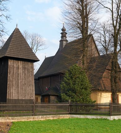 Zdjęcie nr 7: Dzwonnica drewniana, konstrukcji słupowej, ściany szalowane z listowaniem, więźba dachowa drewniana. Założona na planie prostokąta zbliżonego do kwadratu, na betonowym podmurowaniu, przesłoniętym okapowym daszkiem gontowym. Dach gontowy, czterospadowy, zakończony metalowym szpikulcem. Otwór drzwiowy w ścianie południowej o wykroju stojącego prostokąta zamknięty drzwiami spągowymi. Niewielkie otwory głosowe w formie stojącego prostokąta zamkniętego łukiem półkolistym nadwieszonym po dwa na południowej, wschodniej i północnej ścianie. Wnętrze wypełnione elementami konstrukcji.