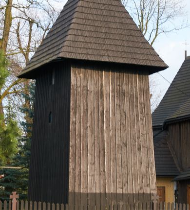 Zdjęcie nr 5: Dzwonnica drewniana, konstrukcji słupowej, ściany szalowane z listowaniem, więźba dachowa drewniana. Założona na planie prostokąta zbliżonego do kwadratu, na betonowym podmurowaniu, przesłoniętym okapowym daszkiem gontowym. Dach gontowy, czterospadowy, zakończony metalowym szpikulcem. Otwór drzwiowy w ścianie południowej o wykroju stojącego prostokąta zamknięty drzwiami spągowymi. Niewielkie otwory głosowe w formie stojącego prostokąta zamkniętego łukiem półkolistym nadwieszonym po dwa na południowej, wschodniej i północnej ścianie. Wnętrze wypełnione elementami konstrukcji.