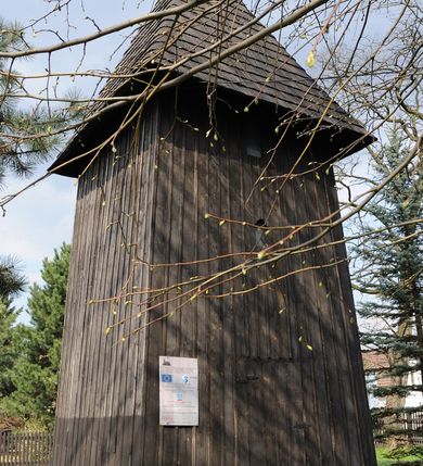 Zdjęcie nr 4: Dzwonnica drewniana, konstrukcji słupowej, ściany szalowane z listowaniem, więźba dachowa drewniana. Założona na planie prostokąta zbliżonego do kwadratu, na betonowym podmurowaniu, przesłoniętym okapowym daszkiem gontowym. Dach gontowy, czterospadowy, zakończony metalowym szpikulcem. Otwór drzwiowy w ścianie południowej o wykroju stojącego prostokąta zamknięty drzwiami spągowymi. Niewielkie otwory głosowe w formie stojącego prostokąta zamkniętego łukiem półkolistym nadwieszonym po dwa na południowej, wschodniej i północnej ścianie. Wnętrze wypełnione elementami konstrukcji.