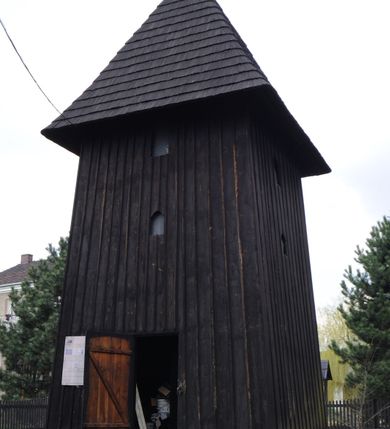 Zdjęcie nr 1: Dzwonnica drewniana, konstrukcji słupowej, ściany szalowane z listowaniem, więźba dachowa drewniana. Założona na planie prostokąta zbliżonego do kwadratu, na betonowym podmurowaniu, przesłoniętym okapowym daszkiem gontowym. Dach gontowy, czterospadowy, zakończony metalowym szpikulcem. Otwór drzwiowy w ścianie południowej o wykroju stojącego prostokąta zamknięty drzwiami spągowymi. Niewielkie otwory głosowe w formie stojącego prostokąta zamkniętego łukiem półkolistym nadwieszonym po dwa na południowej, wschodniej i północnej ścianie. Wnętrze wypełnione elementami konstrukcji.