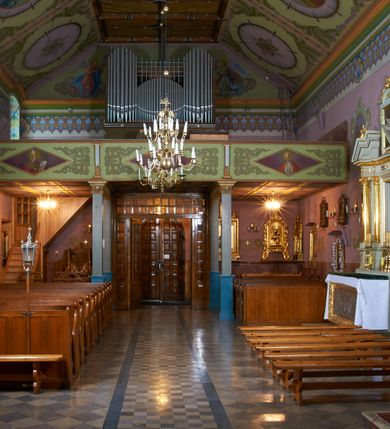 Zdjęcie nr 14: Kościół położony pośrodku wsi, na wysokiej skarpie, wzmocnionej murem. Otoczenie ogrodzone współcześnie. Od wschodu dzwonnica w formie bramy, ze schodami.  Kościół murowany, tynkowany, orientowany, zbudowany na planie wydłużonego prostokąta, jednonawowy, z węższym prezbiterium zamkniętym wielobocznie, opiętym szkarpami. Od północy przylega parterowa zakrystia z przedsionkiem, od południa dwie kruchty. Nawa od prezbiterium oddzielone ostrołukową arkadą. Wnętrze oświetlone oknami prostokątnymi zamkniętymi odcinkowo, wszystkie z witrażami. Nawa nakryta trójspadowym stropem, prezbiterium sklepieniem krzyżowo-żebrowym. Odrzwia z prezbiterium do zakrystii ostrołukowe. Okno w prezbiterium od wschodu ostrołukowe (zamurowane), okno w przedsionku zamknięte tzw. oślim grzbietem. Elewacje na kamiennym, obecnie tynkowanym cokole, oszkarpowane. Szkarpy uskokowe o zmiennym przekroju. Fasada jednoosiowa, zwieńczona trójkątnym szczytem. W osi elewacji ostrołukowy profilowany uskokowo portal, w szczycie okrągły otwór okienny, w płaskiej, tynkowanej opasce. Szczyt dodatkowo podzielony lizenami, u góry zdobiony fryzem arkadkowym, zamknięty wydatnym, profilowanym gzymsem. Oś elewacji zaakcentowana pośrodku wykonanym w tynku i zróżnicowanym kolorystycznie krzyżem, umieszczonym nad wejściem. Po lewej stronie portalu marmurowa tablica epitafijna. Po prawej stronie portalu marmurowa prostokątna tablica z donatorami. Elewacja zachodnia zamknięta wielobocznie, oszkarpowana. W górnej części ostrołukowa blenda, poniżej umieszczony krucyfiks, osłonięty dwuspadowym daszkiem, wspartym na konsolkach. Elewacje boczne pięcioosiowe z prostokątnymi otworami drzwiowymi w drugich osiach od zachodu. Poszczególne osie oddzielone prostokątnymi oknami. Zakrystia i kruchty nakryte dachem jednospadowym. Konstrukcja dachu korpusu głównego drewniana, krokwiowo-płatwiowa. Dach dwuspadowy, nad prezbiterium trójspadowy, kryty blachą ocynkowaną. Sygnaturka w całości obita blachą. Dzwonnica posiada dwie kondygnacje: dolną przeprutą dwiema arkadami, tworzącymi wejście w obręb cmentarza kościelnego i górną, przedzieloną czterema arkadami, przeznaczonymi na pomieszczenie dzwonów. Dach siodłowy.
