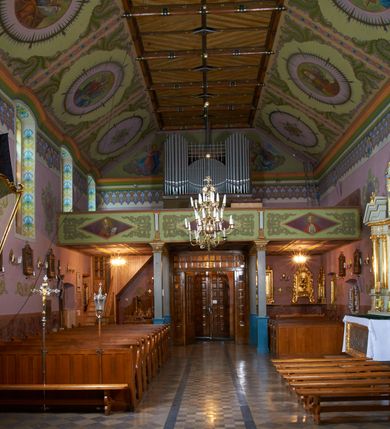 Zdjęcie nr 15: Kościół położony pośrodku wsi, na wysokiej skarpie, wzmocnionej murem. Otoczenie ogrodzone współcześnie. Od wschodu dzwonnica w formie bramy, ze schodami.  Kościół murowany, tynkowany, orientowany, zbudowany na planie wydłużonego prostokąta, jednonawowy, z węższym prezbiterium zamkniętym wielobocznie, opiętym szkarpami. Od północy przylega parterowa zakrystia z przedsionkiem, od południa dwie kruchty. Nawa od prezbiterium oddzielone ostrołukową arkadą. Wnętrze oświetlone oknami prostokątnymi zamkniętymi odcinkowo, wszystkie z witrażami. Nawa nakryta trójspadowym stropem, prezbiterium sklepieniem krzyżowo-żebrowym. Odrzwia z prezbiterium do zakrystii ostrołukowe. Okno w prezbiterium od wschodu ostrołukowe (zamurowane), okno w przedsionku zamknięte tzw. oślim grzbietem. Elewacje na kamiennym, obecnie tynkowanym cokole, oszkarpowane. Szkarpy uskokowe o zmiennym przekroju. Fasada jednoosiowa, zwieńczona trójkątnym szczytem. W osi elewacji ostrołukowy profilowany uskokowo portal, w szczycie okrągły otwór okienny, w płaskiej, tynkowanej opasce. Szczyt dodatkowo podzielony lizenami, u góry zdobiony fryzem arkadkowym, zamknięty wydatnym, profilowanym gzymsem. Oś elewacji zaakcentowana pośrodku wykonanym w tynku i zróżnicowanym kolorystycznie krzyżem, umieszczonym nad wejściem. Po lewej stronie portalu marmurowa tablica epitafijna. Po prawej stronie portalu marmurowa prostokątna tablica z donatorami. Elewacja zachodnia zamknięta wielobocznie, oszkarpowana. W górnej części ostrołukowa blenda, poniżej umieszczony krucyfiks, osłonięty dwuspadowym daszkiem, wspartym na konsolkach. Elewacje boczne pięcioosiowe z prostokątnymi otworami drzwiowymi w drugich osiach od zachodu. Poszczególne osie oddzielone prostokątnymi oknami. Zakrystia i kruchty nakryte dachem jednospadowym. Konstrukcja dachu korpusu głównego drewniana, krokwiowo-płatwiowa. Dach dwuspadowy, nad prezbiterium trójspadowy, kryty blachą ocynkowaną. Sygnaturka w całości obita blachą. Dzwonnica posiada dwie kondygnacje: dolną przeprutą dwiema arkadami, tworzącymi wejście w obręb cmentarza kościelnego i górną, przedzieloną czterema arkadami, przeznaczonymi na pomieszczenie dzwonów. Dach siodłowy.
