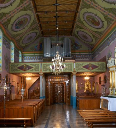 Zdjęcie nr 16: Kościół położony pośrodku wsi, na wysokiej skarpie, wzmocnionej murem. Otoczenie ogrodzone współcześnie. Od wschodu dzwonnica w formie bramy, ze schodami.  Kościół murowany, tynkowany, orientowany, zbudowany na planie wydłużonego prostokąta, jednonawowy, z węższym prezbiterium zamkniętym wielobocznie, opiętym szkarpami. Od północy przylega parterowa zakrystia z przedsionkiem, od południa dwie kruchty. Nawa od prezbiterium oddzielone ostrołukową arkadą. Wnętrze oświetlone oknami prostokątnymi zamkniętymi odcinkowo, wszystkie z witrażami. Nawa nakryta trójspadowym stropem, prezbiterium sklepieniem krzyżowo-żebrowym. Odrzwia z prezbiterium do zakrystii ostrołukowe. Okno w prezbiterium od wschodu ostrołukowe (zamurowane), okno w przedsionku zamknięte tzw. oślim grzbietem. Elewacje na kamiennym, obecnie tynkowanym cokole, oszkarpowane. Szkarpy uskokowe o zmiennym przekroju. Fasada jednoosiowa, zwieńczona trójkątnym szczytem. W osi elewacji ostrołukowy profilowany uskokowo portal, w szczycie okrągły otwór okienny, w płaskiej, tynkowanej opasce. Szczyt dodatkowo podzielony lizenami, u góry zdobiony fryzem arkadkowym, zamknięty wydatnym, profilowanym gzymsem. Oś elewacji zaakcentowana pośrodku wykonanym w tynku i zróżnicowanym kolorystycznie krzyżem, umieszczonym nad wejściem. Po lewej stronie portalu marmurowa tablica epitafijna. Po prawej stronie portalu marmurowa prostokątna tablica z donatorami. Elewacja zachodnia zamknięta wielobocznie, oszkarpowana. W górnej części ostrołukowa blenda, poniżej umieszczony krucyfiks, osłonięty dwuspadowym daszkiem, wspartym na konsolkach. Elewacje boczne pięcioosiowe z prostokątnymi otworami drzwiowymi w drugich osiach od zachodu. Poszczególne osie oddzielone prostokątnymi oknami. Zakrystia i kruchty nakryte dachem jednospadowym. Konstrukcja dachu korpusu głównego drewniana, krokwiowo-płatwiowa. Dach dwuspadowy, nad prezbiterium trójspadowy, kryty blachą ocynkowaną. Sygnaturka w całości obita blachą. Dzwonnica posiada dwie kondygnacje: dolną przeprutą dwiema arkadami, tworzącymi wejście w obręb cmentarza kościelnego i górną, przedzieloną czterema arkadami, przeznaczonymi na pomieszczenie dzwonów. Dach siodłowy.
