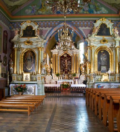 Zdjęcie nr 11: Kościół położony pośrodku wsi, na wysokiej skarpie, wzmocnionej murem. Otoczenie ogrodzone współcześnie. Od wschodu dzwonnica w formie bramy, ze schodami.  Kościół murowany, tynkowany, orientowany, zbudowany na planie wydłużonego prostokąta, jednonawowy, z węższym prezbiterium zamkniętym wielobocznie, opiętym szkarpami. Od północy przylega parterowa zakrystia z przedsionkiem, od południa dwie kruchty. Nawa od prezbiterium oddzielone ostrołukową arkadą. Wnętrze oświetlone oknami prostokątnymi zamkniętymi odcinkowo, wszystkie z witrażami. Nawa nakryta trójspadowym stropem, prezbiterium sklepieniem krzyżowo-żebrowym. Odrzwia z prezbiterium do zakrystii ostrołukowe. Okno w prezbiterium od wschodu ostrołukowe (zamurowane), okno w przedsionku zamknięte tzw. oślim grzbietem. Elewacje na kamiennym, obecnie tynkowanym cokole, oszkarpowane. Szkarpy uskokowe o zmiennym przekroju. Fasada jednoosiowa, zwieńczona trójkątnym szczytem. W osi elewacji ostrołukowy profilowany uskokowo portal, w szczycie okrągły otwór okienny, w płaskiej, tynkowanej opasce. Szczyt dodatkowo podzielony lizenami, u góry zdobiony fryzem arkadkowym, zamknięty wydatnym, profilowanym gzymsem. Oś elewacji zaakcentowana pośrodku wykonanym w tynku i zróżnicowanym kolorystycznie krzyżem, umieszczonym nad wejściem. Po lewej stronie portalu marmurowa tablica epitafijna. Po prawej stronie portalu marmurowa prostokątna tablica z donatorami. Elewacja zachodnia zamknięta wielobocznie, oszkarpowana. W górnej części ostrołukowa blenda, poniżej umieszczony krucyfiks, osłonięty dwuspadowym daszkiem, wspartym na konsolkach. Elewacje boczne pięcioosiowe z prostokątnymi otworami drzwiowymi w drugich osiach od zachodu. Poszczególne osie oddzielone prostokątnymi oknami. Zakrystia i kruchty nakryte dachem jednospadowym. Konstrukcja dachu korpusu głównego drewniana, krokwiowo-płatwiowa. Dach dwuspadowy, nad prezbiterium trójspadowy, kryty blachą ocynkowaną. Sygnaturka w całości obita blachą. Dzwonnica posiada dwie kondygnacje: dolną przeprutą dwiema arkadami, tworzącymi wejście w obręb cmentarza kościelnego i górną, przedzieloną czterema arkadami, przeznaczonymi na pomieszczenie dzwonów. Dach siodłowy.
