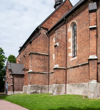 Zdjęcie nr 31: Kościół jest zlokalizowany na wzgórzu, w centrum wsi. Murowany z cegły, w wątku polskim, z zendrówką. Kościół jest orientowany, założony na planie prostokąta, jednonawowy, trójprzęsłowy, z dobudowanymi kruchtami od zachodu i południa. Prezbiterium oddzielone od nawy łukiem tęczowym, dwuprzęsłowe na planie prostokąta, zamknięte prostą ścianą, z dwukondygnacyjną przybudówką zakończoną trójbocznie. Nawa została przykryta sklepieniem sieciowym. Ściana południowa nawy przepruta trzema prostokątnymi oknami, zamkniętymi łukiem ostrym. Okna w nawie i prezbiterium dwudzielne, z sześcioma mniejszymi kwaterami. Przybudówka przy prezbiterium na parterze mieści zakrystię, a na piętrze składzik. Kruchta południowa sklepiona kolebką. Z kruchty do nawy prowadzi ostrołukowy, kamienny portal o dwóch archiwoltach. Kruchta zachodnia, obecnie kaplica św. Franciszka, sklepiona spłaszczoną, ukrytą kopułą na pendentywach. Ściana zachodnia kruchty przepruta dwoma ostrołukowymi oknami i ponad nimi okrągłym oknem. Od południa ostrołukowe wejście. Kościół o zróżnicowanej wysokości nawy i prezbiterium. Ściany opięte szkarpami. Na jednej trzeciej wysokości ścian oraz w zwieńczeniu nawy i prezbiterium profilowany gzyms. Od zachodu wklęsło-wypukły szczyt. Nawa, prezbiterium i dwie kruchty przykryte dachem dwuspadowym. Nad drugim przęsłem prezbiterium wieżyczka na sygnaturkę na planie ośmioboku, z profilowanym gzymsem i latarnią, zwieńczoną hełmem z kulą i krzyżem. Wejście do kościoła od południa i zachodu. Okna nawy i prezbiterium z kamiennym obramieniem. Nad wejściem do kruchty południowej formowany w cegle ostry łuk. Analogiczny łuk nad oknami w ścianie zachodniej i nad blendą w ścianie północnej. W przybudówce do prezbiterium dwie kondygnacje okien zamkniętych łukiem ostrym. W ścianie frontowej prostokątne wejście, a nad nim półkoliście zamknięta wnęka z grupą Ukrzyżowania. 


