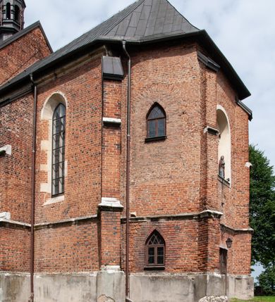 Zdjęcie nr 27: Kościół jest zlokalizowany na wzgórzu, w centrum wsi. Murowany z cegły, w wątku polskim, z zendrówką. Kościół jest orientowany, założony na planie prostokąta, jednonawowy, trójprzęsłowy, z dobudowanymi kruchtami od zachodu i południa. Prezbiterium oddzielone od nawy łukiem tęczowym, dwuprzęsłowe na planie prostokąta, zamknięte prostą ścianą, z dwukondygnacyjną przybudówką zakończoną trójbocznie. Nawa została przykryta sklepieniem sieciowym. Ściana południowa nawy przepruta trzema prostokątnymi oknami, zamkniętymi łukiem ostrym. Okna w nawie i prezbiterium dwudzielne, z sześcioma mniejszymi kwaterami. Przybudówka przy prezbiterium na parterze mieści zakrystię, a na piętrze składzik. Kruchta południowa sklepiona kolebką. Z kruchty do nawy prowadzi ostrołukowy, kamienny portal o dwóch archiwoltach. Kruchta zachodnia, obecnie kaplica św. Franciszka, sklepiona spłaszczoną, ukrytą kopułą na pendentywach. Ściana zachodnia kruchty przepruta dwoma ostrołukowymi oknami i ponad nimi okrągłym oknem. Od południa ostrołukowe wejście. Kościół o zróżnicowanej wysokości nawy i prezbiterium. Ściany opięte szkarpami. Na jednej trzeciej wysokości ścian oraz w zwieńczeniu nawy i prezbiterium profilowany gzyms. Od zachodu wklęsło-wypukły szczyt. Nawa, prezbiterium i dwie kruchty przykryte dachem dwuspadowym. Nad drugim przęsłem prezbiterium wieżyczka na sygnaturkę na planie ośmioboku, z profilowanym gzymsem i latarnią, zwieńczoną hełmem z kulą i krzyżem. Wejście do kościoła od południa i zachodu. Okna nawy i prezbiterium z kamiennym obramieniem. Nad wejściem do kruchty południowej formowany w cegle ostry łuk. Analogiczny łuk nad oknami w ścianie zachodniej i nad blendą w ścianie północnej. W przybudówce do prezbiterium dwie kondygnacje okien zamkniętych łukiem ostrym. W ścianie frontowej prostokątne wejście, a nad nim półkoliście zamknięta wnęka z grupą Ukrzyżowania. 


