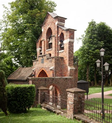 Zdjęcie nr 12: Kościół jest zlokalizowany na wzgórzu, w centrum wsi. Murowany z cegły, w wątku polskim, z zendrówką. Kościół jest orientowany, założony na planie prostokąta, jednonawowy, trójprzęsłowy, z dobudowanymi kruchtami od zachodu i południa. Prezbiterium oddzielone od nawy łukiem tęczowym, dwuprzęsłowe na planie prostokąta, zamknięte prostą ścianą, z dwukondygnacyjną przybudówką zakończoną trójbocznie. Nawa została przykryta sklepieniem sieciowym. Ściana południowa nawy przepruta trzema prostokątnymi oknami, zamkniętymi łukiem ostrym. Okna w nawie i prezbiterium dwudzielne, z sześcioma mniejszymi kwaterami. Przybudówka przy prezbiterium na parterze mieści zakrystię, a na piętrze składzik. Kruchta południowa sklepiona kolebką. Z kruchty do nawy prowadzi ostrołukowy, kamienny portal o dwóch archiwoltach. Kruchta zachodnia, obecnie kaplica św. Franciszka, sklepiona spłaszczoną, ukrytą kopułą na pendentywach. Ściana zachodnia kruchty przepruta dwoma ostrołukowymi oknami i ponad nimi okrągłym oknem. Od południa ostrołukowe wejście. Kościół o zróżnicowanej wysokości nawy i prezbiterium. Ściany opięte szkarpami. Na jednej trzeciej wysokości ścian oraz w zwieńczeniu nawy i prezbiterium profilowany gzyms. Od zachodu wklęsło-wypukły szczyt. Nawa, prezbiterium i dwie kruchty przykryte dachem dwuspadowym. Nad drugim przęsłem prezbiterium wieżyczka na sygnaturkę na planie ośmioboku, z profilowanym gzymsem i latarnią, zwieńczoną hełmem z kulą i krzyżem. Wejście do kościoła od południa i zachodu. Okna nawy i prezbiterium z kamiennym obramieniem. Nad wejściem do kruchty południowej formowany w cegle ostry łuk. Analogiczny łuk nad oknami w ścianie zachodniej i nad blendą w ścianie północnej. W przybudówce do prezbiterium dwie kondygnacje okien zamkniętych łukiem ostrym. W ścianie frontowej prostokątne wejście, a nad nim półkoliście zamknięta wnęka z grupą Ukrzyżowania. 


