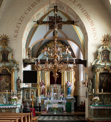 Zdjęcie nr 89: Kościół jest zlokalizowany na wzgórzu, w centrum wsi. Murowany z cegły, w wątku polskim, z zendrówką. Kościół jest orientowany, założony na planie prostokąta, jednonawowy, trójprzęsłowy, z dobudowanymi kruchtami od zachodu i południa. Prezbiterium oddzielone od nawy łukiem tęczowym, dwuprzęsłowe na planie prostokąta, zamknięte prostą ścianą, z dwukondygnacyjną przybudówką zakończoną trójbocznie. Nawa została przykryta sklepieniem sieciowym. Ściana południowa nawy przepruta trzema prostokątnymi oknami, zamkniętymi łukiem ostrym. Okna w nawie i prezbiterium dwudzielne, z sześcioma mniejszymi kwaterami. Przybudówka przy prezbiterium na parterze mieści zakrystię, a na piętrze składzik. Kruchta południowa sklepiona kolebką. Z kruchty do nawy prowadzi ostrołukowy, kamienny portal o dwóch archiwoltach. Kruchta zachodnia, obecnie kaplica św. Franciszka, sklepiona spłaszczoną, ukrytą kopułą na pendentywach. Ściana zachodnia kruchty przepruta dwoma ostrołukowymi oknami i ponad nimi okrągłym oknem. Od południa ostrołukowe wejście. Kościół o zróżnicowanej wysokości nawy i prezbiterium. Ściany opięte szkarpami. Na jednej trzeciej wysokości ścian oraz w zwieńczeniu nawy i prezbiterium profilowany gzyms. Od zachodu wklęsło-wypukły szczyt. Nawa, prezbiterium i dwie kruchty przykryte dachem dwuspadowym. Nad drugim przęsłem prezbiterium wieżyczka na sygnaturkę na planie ośmioboku, z profilowanym gzymsem i latarnią, zwieńczoną hełmem z kulą i krzyżem. Wejście do kościoła od południa i zachodu. Okna nawy i prezbiterium z kamiennym obramieniem. Nad wejściem do kruchty południowej formowany w cegle ostry łuk. Analogiczny łuk nad oknami w ścianie zachodniej i nad blendą w ścianie północnej. W przybudówce do prezbiterium dwie kondygnacje okien zamkniętych łukiem ostrym. W ścianie frontowej prostokątne wejście, a nad nim półkoliście zamknięta wnęka z grupą Ukrzyżowania. 


