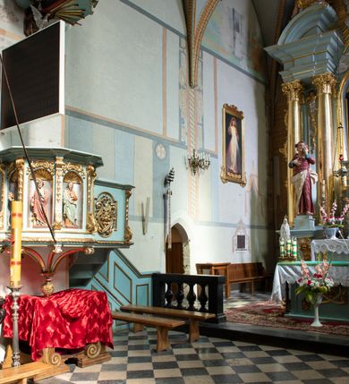 Zdjęcie nr 94: Kościół jest zlokalizowany na wzgórzu, w centrum wsi. Murowany z cegły, w wątku polskim, z zendrówką. Kościół jest orientowany, założony na planie prostokąta, jednonawowy, trójprzęsłowy, z dobudowanymi kruchtami od zachodu i południa. Prezbiterium oddzielone od nawy łukiem tęczowym, dwuprzęsłowe na planie prostokąta, zamknięte prostą ścianą, z dwukondygnacyjną przybudówką zakończoną trójbocznie. Nawa została przykryta sklepieniem sieciowym. Ściana południowa nawy przepruta trzema prostokątnymi oknami, zamkniętymi łukiem ostrym. Okna w nawie i prezbiterium dwudzielne, z sześcioma mniejszymi kwaterami. Przybudówka przy prezbiterium na parterze mieści zakrystię, a na piętrze składzik. Kruchta południowa sklepiona kolebką. Z kruchty do nawy prowadzi ostrołukowy, kamienny portal o dwóch archiwoltach. Kruchta zachodnia, obecnie kaplica św. Franciszka, sklepiona spłaszczoną, ukrytą kopułą na pendentywach. Ściana zachodnia kruchty przepruta dwoma ostrołukowymi oknami i ponad nimi okrągłym oknem. Od południa ostrołukowe wejście. Kościół o zróżnicowanej wysokości nawy i prezbiterium. Ściany opięte szkarpami. Na jednej trzeciej wysokości ścian oraz w zwieńczeniu nawy i prezbiterium profilowany gzyms. Od zachodu wklęsło-wypukły szczyt. Nawa, prezbiterium i dwie kruchty przykryte dachem dwuspadowym. Nad drugim przęsłem prezbiterium wieżyczka na sygnaturkę na planie ośmioboku, z profilowanym gzymsem i latarnią, zwieńczoną hełmem z kulą i krzyżem. Wejście do kościoła od południa i zachodu. Okna nawy i prezbiterium z kamiennym obramieniem. Nad wejściem do kruchty południowej formowany w cegle ostry łuk. Analogiczny łuk nad oknami w ścianie zachodniej i nad blendą w ścianie północnej. W przybudówce do prezbiterium dwie kondygnacje okien zamkniętych łukiem ostrym. W ścianie frontowej prostokątne wejście, a nad nim półkoliście zamknięta wnęka z grupą Ukrzyżowania. 


