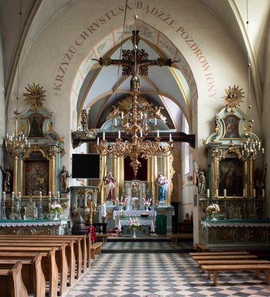 Zdjęcie nr 92: Kościół jest zlokalizowany na wzgórzu, w centrum wsi. Murowany z cegły, w wątku polskim, z zendrówką. Kościół jest orientowany, założony na planie prostokąta, jednonawowy, trójprzęsłowy, z dobudowanymi kruchtami od zachodu i południa. Prezbiterium oddzielone od nawy łukiem tęczowym, dwuprzęsłowe na planie prostokąta, zamknięte prostą ścianą, z dwukondygnacyjną przybudówką zakończoną trójbocznie. Nawa została przykryta sklepieniem sieciowym. Ściana południowa nawy przepruta trzema prostokątnymi oknami, zamkniętymi łukiem ostrym. Okna w nawie i prezbiterium dwudzielne, z sześcioma mniejszymi kwaterami. Przybudówka przy prezbiterium na parterze mieści zakrystię, a na piętrze składzik. Kruchta południowa sklepiona kolebką. Z kruchty do nawy prowadzi ostrołukowy, kamienny portal o dwóch archiwoltach. Kruchta zachodnia, obecnie kaplica św. Franciszka, sklepiona spłaszczoną, ukrytą kopułą na pendentywach. Ściana zachodnia kruchty przepruta dwoma ostrołukowymi oknami i ponad nimi okrągłym oknem. Od południa ostrołukowe wejście. Kościół o zróżnicowanej wysokości nawy i prezbiterium. Ściany opięte szkarpami. Na jednej trzeciej wysokości ścian oraz w zwieńczeniu nawy i prezbiterium profilowany gzyms. Od zachodu wklęsło-wypukły szczyt. Nawa, prezbiterium i dwie kruchty przykryte dachem dwuspadowym. Nad drugim przęsłem prezbiterium wieżyczka na sygnaturkę na planie ośmioboku, z profilowanym gzymsem i latarnią, zwieńczoną hełmem z kulą i krzyżem. Wejście do kościoła od południa i zachodu. Okna nawy i prezbiterium z kamiennym obramieniem. Nad wejściem do kruchty południowej formowany w cegle ostry łuk. Analogiczny łuk nad oknami w ścianie zachodniej i nad blendą w ścianie północnej. W przybudówce do prezbiterium dwie kondygnacje okien zamkniętych łukiem ostrym. W ścianie frontowej prostokątne wejście, a nad nim półkoliście zamknięta wnęka z grupą Ukrzyżowania. 


