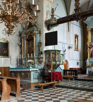 Zdjęcie nr 97: Kościół jest zlokalizowany na wzgórzu, w centrum wsi. Murowany z cegły, w wątku polskim, z zendrówką. Kościół jest orientowany, założony na planie prostokąta, jednonawowy, trójprzęsłowy, z dobudowanymi kruchtami od zachodu i południa. Prezbiterium oddzielone od nawy łukiem tęczowym, dwuprzęsłowe na planie prostokąta, zamknięte prostą ścianą, z dwukondygnacyjną przybudówką zakończoną trójbocznie. Nawa została przykryta sklepieniem sieciowym. Ściana południowa nawy przepruta trzema prostokątnymi oknami, zamkniętymi łukiem ostrym. Okna w nawie i prezbiterium dwudzielne, z sześcioma mniejszymi kwaterami. Przybudówka przy prezbiterium na parterze mieści zakrystię, a na piętrze składzik. Kruchta południowa sklepiona kolebką. Z kruchty do nawy prowadzi ostrołukowy, kamienny portal o dwóch archiwoltach. Kruchta zachodnia, obecnie kaplica św. Franciszka, sklepiona spłaszczoną, ukrytą kopułą na pendentywach. Ściana zachodnia kruchty przepruta dwoma ostrołukowymi oknami i ponad nimi okrągłym oknem. Od południa ostrołukowe wejście. Kościół o zróżnicowanej wysokości nawy i prezbiterium. Ściany opięte szkarpami. Na jednej trzeciej wysokości ścian oraz w zwieńczeniu nawy i prezbiterium profilowany gzyms. Od zachodu wklęsło-wypukły szczyt. Nawa, prezbiterium i dwie kruchty przykryte dachem dwuspadowym. Nad drugim przęsłem prezbiterium wieżyczka na sygnaturkę na planie ośmioboku, z profilowanym gzymsem i latarnią, zwieńczoną hełmem z kulą i krzyżem. Wejście do kościoła od południa i zachodu. Okna nawy i prezbiterium z kamiennym obramieniem. Nad wejściem do kruchty południowej formowany w cegle ostry łuk. Analogiczny łuk nad oknami w ścianie zachodniej i nad blendą w ścianie północnej. W przybudówce do prezbiterium dwie kondygnacje okien zamkniętych łukiem ostrym. W ścianie frontowej prostokątne wejście, a nad nim półkoliście zamknięta wnęka z grupą Ukrzyżowania. 


