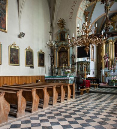 Zdjęcie nr 99: Kościół jest zlokalizowany na wzgórzu, w centrum wsi. Murowany z cegły, w wątku polskim, z zendrówką. Kościół jest orientowany, założony na planie prostokąta, jednonawowy, trójprzęsłowy, z dobudowanymi kruchtami od zachodu i południa. Prezbiterium oddzielone od nawy łukiem tęczowym, dwuprzęsłowe na planie prostokąta, zamknięte prostą ścianą, z dwukondygnacyjną przybudówką zakończoną trójbocznie. Nawa została przykryta sklepieniem sieciowym. Ściana południowa nawy przepruta trzema prostokątnymi oknami, zamkniętymi łukiem ostrym. Okna w nawie i prezbiterium dwudzielne, z sześcioma mniejszymi kwaterami. Przybudówka przy prezbiterium na parterze mieści zakrystię, a na piętrze składzik. Kruchta południowa sklepiona kolebką. Z kruchty do nawy prowadzi ostrołukowy, kamienny portal o dwóch archiwoltach. Kruchta zachodnia, obecnie kaplica św. Franciszka, sklepiona spłaszczoną, ukrytą kopułą na pendentywach. Ściana zachodnia kruchty przepruta dwoma ostrołukowymi oknami i ponad nimi okrągłym oknem. Od południa ostrołukowe wejście. Kościół o zróżnicowanej wysokości nawy i prezbiterium. Ściany opięte szkarpami. Na jednej trzeciej wysokości ścian oraz w zwieńczeniu nawy i prezbiterium profilowany gzyms. Od zachodu wklęsło-wypukły szczyt. Nawa, prezbiterium i dwie kruchty przykryte dachem dwuspadowym. Nad drugim przęsłem prezbiterium wieżyczka na sygnaturkę na planie ośmioboku, z profilowanym gzymsem i latarnią, zwieńczoną hełmem z kulą i krzyżem. Wejście do kościoła od południa i zachodu. Okna nawy i prezbiterium z kamiennym obramieniem. Nad wejściem do kruchty południowej formowany w cegle ostry łuk. Analogiczny łuk nad oknami w ścianie zachodniej i nad blendą w ścianie północnej. W przybudówce do prezbiterium dwie kondygnacje okien zamkniętych łukiem ostrym. W ścianie frontowej prostokątne wejście, a nad nim półkoliście zamknięta wnęka z grupą Ukrzyżowania. 


