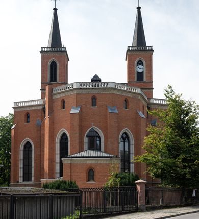 Zdjęcie nr 4: Kościół z cegły i kamienia. Na rzucie krzyża łacińskiego, jednonawowy z wciągniętymi do wnętrza przyporami. Prezbiterium krótkie, zamknięte pięcioma bokami ośmioboku. Korpus jednonawowy, czteroprzęsłowy, po bokach z płytkimi wnękami-kaplicami pomiędzy przyporami, poprzedzony od zach. przedsionkiem. Do pierwszego przęsła dostawione dwie symetryczne, trójboczne kaplice, nad nimi dwie czworoboczne wieże. Zakrystia przy prezbiterium na osi kościoła. Artykulacja ścian wnętrza za pomocą filarów opiętych półkolumnami z kapitelami z motywów liści. Między filarami rozpięte ostrołukowe arkady tworzące płytkie wnęki. Pierwsze przęsło wyróżnione przy pomocy masywnych filarów, ujętych niemal pełnoplastycznymi kolumnami. Okna ostrołukowe, dwudzielne z maswerkami, wsparte na gzymsach. Sklepienia krzyżowo-żebrowe na zaostrzonych gurtach. Kaplice artykułowane analogicznie do nawy kościoła, ze sklepieniami gwiaździstymi. Przedsionek otwiera się do kościoła trzema portalami ostrołukowymi opiętymi kolumnami. W polach archiwolt płaskorzeźby: Boże Narodzenie, Ukrzyżowanie, Wniebowstąpienie (K. Ceptowski).  W prezbiterium, na osi kościoła prostokątny otwór drzwiowy prowadzący do zakrystii. Chór muzyczny umieszczony nad kruchtą kościoła, na rzucie prostokąta, z kamienną balustradą maswerkową. Fasada kamienna, trójosiowa, zamknięta trójkątnie, flankowana lizenami przechodzącymi w ażurowe wieżyczki-latarnie, na rzucie kwadratu. Wieżyczki zwieńczone gzymsem i balustradą arkadową, hełmy ostrosłupowy zakończony iglicą. W dolnej części trzy filarowe arkady ostrołukowe, otwierające się do kruchty, sklepienie kruchty krzyżowo-żebrowe. Nad arkadami wejściowymi trzy tonda: litery IHS, Oko Opatrzności i splecione litery imienia Maria. Pośrodku na osi kościoła duża rozeta z maswerkiem, profilowana, otoczona pierścieniem wypełnionym stylizowanym ornamentem roślinnym. Ponad arkadami pełnoplastyczne figury czterech ewangelistów ustawionych na konsolach z rzeźbionymi atrybutami (F. Kuhn). Po bokach rozety prostokątne płyciny z traczami herbowymi Potockich i Branickich. U góry, na osi fasady figura Archanioła Michała. Elewacje na kamiennym cokole, z gzymsem kapnikowym, opięte prostokątnymi przyporami zwieńczonymi kamiennymi nakrywami. W górnej części kamienny fryz arkadkowy. Nad nim gzyms koronujący podtrzymujący ażurową, arkadową balustradę. Przy prezbiterium kaplice zamknięte trójbocznie, prezbiterium zamknięte pięciobocznie. Zakrystia na osi prezbiterium, na rzucie prostokąta, zwieńczona sygnaturką i ceglanym kominem. Po bokach prostokątne aneksy, południowy aneks - trzy okna ostrołukowe w kamiennej opasce, w środkowym przedstawienie Ukrzyżowanego Chrystusa. W północnym aneksie pojedyncze analogiczne okno. Okna ostrołukowe w profilowanych kamiennych opaskach, pomiędzy nimi a gzymsem koronującym mniejsze ostrołukowe w uskokowych arkadach. Nad kaplicami bocznymi dwie symetryczne wieże na rzucie kwadratu, w połowie wysokości kamienny gzyms z fryzem arkadkowym. Okna ostrołukowe w kamiennej opasce. Całość zwieńczona ślepym fryzem arkadkowym, wyżej gzyms dźwigający ażurową arkadkową balustradę. Hełmy ostrosłupowe, miedziane. Zakrystia na rzucie prostokąta, nakryta sklepieniem kolebkowym zaostrzonym i krzyżowym z lunetami , oddzielone gurtem. Portale od wschodu i zachodu prostokątne zamknięte łukiem ostrym. Okna ostrołukowe.