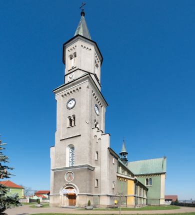 Zdjęcie nr 1: Budynki kościoła i plebanii znajdują się w centralnej części wsi, przy rynku na północ od głównej drogi z Krakowa do Oświęcimia. Kościół stoi przy obszernym placu, obwiedziony wysokim kamiennym murem z metalowym ogrodzeniem od południa. Kościół z prezbiterium zwróconym na północ, wzniesiony z kamienia wapiennego i cegły, tynkowany, z podmurówką. Świątynia wybudowana na planie krzyża łacińskiego z bazylikowym korpusem i transeptem. Dwuprzęsłowe prezbiterium zakończone półkoliście absydą zostało ujęte w dwa aneksy boczne, z których zachodni przeznaczony jest na zakrystię. Od południa do budynku przylega kruchta w przyziemiu wieży. Wnętrze kościoła trójnawowe, czteroprzęsłowe. Nawa główna łączy się z nawami bocznymi arkadami wspartymi na pięciobocznych filarach. Nawy boczne ujęte szerokimi pilastrami z pięciobocznymi filarami. Filary i pilastry stoją na wysokich, masywnych cokołach z kapitelami analogicznymi jak w nawie głównej. Ściany na skrzyżowaniu naw oraz ściany prezbiterium ujęte wspomnianymi filarami podtrzymującymi gurty sklepienia. Nawa i prezbiterium przykryte są sklepieniem kolebkowym z geometryzowanymi lunetami po obu stronach ścian. Ramiona transeptu sklepione kolebkowo z analogicznymi lunetami. Na skrzyżowaniu naw znajduje się sklepienie żaglaste. Nawę główną oświetlają rozmieszczone po jednym w każdym przęśle otwory okienne zamknięte łukiem półkolistym, okna w transepcie to biforia, widoczne tylko z zewnątrz okrągłe okno w absydzie wypełnione jest kamienną rozetą. Główne wejście od południa, do pierwszego przęsła od transeptu prowadzi dwoje drzwi w otworach o zamknięciu w kształcie łuku odciętego. W części zachodniej chór muzyczny wsparty na dwóch kolumnach i dwóch filarach dostawionych do ściany. Ich kapitele są analogiczne do pozostałych w kościele. Sklepienia dekorowane polichromią z rozetami i pękami lilii, a na skrzyżowaniu naw umieszczono malunek naścienny ukazujący Chrystusa tronującego z Bogiem Ojcem i gołębicą Ducha Świętego, a wokół nich cztery symbole ewangelistów. W fasadzie świątyni znajduje się strzelista wieża dwukondygnacyjna na planie czworokąta, przechodząca wyżej w ośmioboczną. Po bokach wieży wysokie aneksy zwieńczone schodkowo. Nad wejściem do kościoła znajduje się ozdobne okno zamknięte półkoliście i wypełnione czterema małymi rozetami ułożonymi jedna nad drugą. W kolejnej kondygnacji wieżę przepruwa biforium poniżej tarczy zegara i dekoracji arkadkowej. Wieża pełni jednocześnie funkcję dzwonnicy. Druga wieża na skrzyżowaniu nawy głównej i transeptu mieści sygnaturkę. Korpus kościoła nakryty dachem dwuspadowym, wieża wielobocznym. Wszystkie dachy kryte blachą.