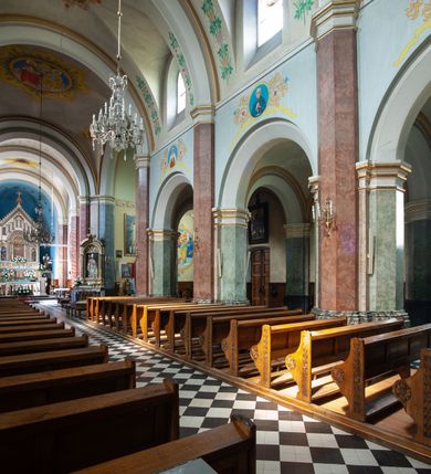 Zdjęcie nr 34: Budynki kościoła i plebanii znajdują się w centralnej części wsi, przy rynku na północ od głównej drogi z Krakowa do Oświęcimia. Kościół stoi przy obszernym placu, obwiedziony wysokim kamiennym murem z metalowym ogrodzeniem od południa. Kościół z prezbiterium zwróconym na północ, wzniesiony z kamienia wapiennego i cegły, tynkowany, z podmurówką. Świątynia wybudowana na planie krzyża łacińskiego z bazylikowym korpusem i transeptem. Dwuprzęsłowe prezbiterium zakończone półkoliście absydą zostało ujęte w dwa aneksy boczne, z których zachodni przeznaczony jest na zakrystię. Od południa do budynku przylega kruchta w przyziemiu wieży. Wnętrze kościoła trójnawowe, czteroprzęsłowe. Nawa główna łączy się z nawami bocznymi arkadami wspartymi na pięciobocznych filarach. Nawy boczne ujęte szerokimi pilastrami z pięciobocznymi filarami. Filary i pilastry stoją na wysokich, masywnych cokołach z kapitelami analogicznymi jak w nawie głównej. Ściany na skrzyżowaniu naw oraz ściany prezbiterium ujęte wspomnianymi filarami podtrzymującymi gurty sklepienia. Nawa i prezbiterium przykryte są sklepieniem kolebkowym z geometryzowanymi lunetami po obu stronach ścian. Ramiona transeptu sklepione kolebkowo z analogicznymi lunetami. Na skrzyżowaniu naw znajduje się sklepienie żaglaste. Nawę główną oświetlają rozmieszczone po jednym w każdym przęśle otwory okienne zamknięte łukiem półkolistym, okna w transepcie to biforia, widoczne tylko z zewnątrz okrągłe okno w absydzie wypełnione jest kamienną rozetą. Główne wejście od południa, do pierwszego przęsła od transeptu prowadzi dwoje drzwi w otworach o zamknięciu w kształcie łuku odciętego. W części zachodniej chór muzyczny wsparty na dwóch kolumnach i dwóch filarach dostawionych do ściany. Ich kapitele są analogiczne do pozostałych w kościele. Sklepienia dekorowane polichromią z rozetami i pękami lilii, a na skrzyżowaniu naw umieszczono malunek naścienny ukazujący Chrystusa tronującego z Bogiem Ojcem i gołębicą Ducha Świętego, a wokół nich cztery symbole ewangelistów. W fasadzie świątyni znajduje się strzelista wieża dwukondygnacyjna na planie czworokąta, przechodząca wyżej w ośmioboczną. Po bokach wieży wysokie aneksy zwieńczone schodkowo. Nad wejściem do kościoła znajduje się ozdobne okno zamknięte półkoliście i wypełnione czterema małymi rozetami ułożonymi jedna nad drugą. W kolejnej kondygnacji wieżę przepruwa biforium poniżej tarczy zegara i dekoracji arkadkowej. Wieża pełni jednocześnie funkcję dzwonnicy. Druga wieża na skrzyżowaniu nawy głównej i transeptu mieści sygnaturkę. Korpus kościoła nakryty dachem dwuspadowym, wieża wielobocznym. Wszystkie dachy kryte blachą.