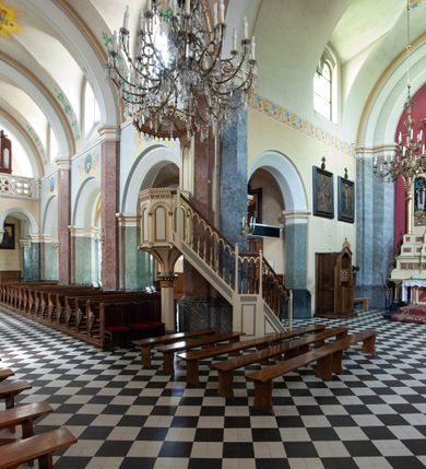 Zdjęcie nr 41: Budynki kościoła i plebanii znajdują się w centralnej części wsi, przy rynku na północ od głównej drogi z Krakowa do Oświęcimia. Kościół stoi przy obszernym placu, obwiedziony wysokim kamiennym murem z metalowym ogrodzeniem od południa. Kościół z prezbiterium zwróconym na północ, wzniesiony z kamienia wapiennego i cegły, tynkowany, z podmurówką. Świątynia wybudowana na planie krzyża łacińskiego z bazylikowym korpusem i transeptem. Dwuprzęsłowe prezbiterium zakończone półkoliście absydą zostało ujęte w dwa aneksy boczne, z których zachodni przeznaczony jest na zakrystię. Od południa do budynku przylega kruchta w przyziemiu wieży. Wnętrze kościoła trójnawowe, czteroprzęsłowe. Nawa główna łączy się z nawami bocznymi arkadami wspartymi na pięciobocznych filarach. Nawy boczne ujęte szerokimi pilastrami z pięciobocznymi filarami. Filary i pilastry stoją na wysokich, masywnych cokołach z kapitelami analogicznymi jak w nawie głównej. Ściany na skrzyżowaniu naw oraz ściany prezbiterium ujęte wspomnianymi filarami podtrzymującymi gurty sklepienia. Nawa i prezbiterium przykryte są sklepieniem kolebkowym z geometryzowanymi lunetami po obu stronach ścian. Ramiona transeptu sklepione kolebkowo z analogicznymi lunetami. Na skrzyżowaniu naw znajduje się sklepienie żaglaste. Nawę główną oświetlają rozmieszczone po jednym w każdym przęśle otwory okienne zamknięte łukiem półkolistym, okna w transepcie to biforia, widoczne tylko z zewnątrz okrągłe okno w absydzie wypełnione jest kamienną rozetą. Główne wejście od południa, do pierwszego przęsła od transeptu prowadzi dwoje drzwi w otworach o zamknięciu w kształcie łuku odciętego. W części zachodniej chór muzyczny wsparty na dwóch kolumnach i dwóch filarach dostawionych do ściany. Ich kapitele są analogiczne do pozostałych w kościele. Sklepienia dekorowane polichromią z rozetami i pękami lilii, a na skrzyżowaniu naw umieszczono malunek naścienny ukazujący Chrystusa tronującego z Bogiem Ojcem i gołębicą Ducha Świętego, a wokół nich cztery symbole ewangelistów. W fasadzie świątyni znajduje się strzelista wieża dwukondygnacyjna na planie czworokąta, przechodząca wyżej w ośmioboczną. Po bokach wieży wysokie aneksy zwieńczone schodkowo. Nad wejściem do kościoła znajduje się ozdobne okno zamknięte półkoliście i wypełnione czterema małymi rozetami ułożonymi jedna nad drugą. W kolejnej kondygnacji wieżę przepruwa biforium poniżej tarczy zegara i dekoracji arkadkowej. Wieża pełni jednocześnie funkcję dzwonnicy. Druga wieża na skrzyżowaniu nawy głównej i transeptu mieści sygnaturkę. Korpus kościoła nakryty dachem dwuspadowym, wieża wielobocznym. Wszystkie dachy kryte blachą.