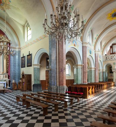 Zdjęcie nr 40: Budynki kościoła i plebanii znajdują się w centralnej części wsi, przy rynku na północ od głównej drogi z Krakowa do Oświęcimia. Kościół stoi przy obszernym placu, obwiedziony wysokim kamiennym murem z metalowym ogrodzeniem od południa. Kościół z prezbiterium zwróconym na północ, wzniesiony z kamienia wapiennego i cegły, tynkowany, z podmurówką. Świątynia wybudowana na planie krzyża łacińskiego z bazylikowym korpusem i transeptem. Dwuprzęsłowe prezbiterium zakończone półkoliście absydą zostało ujęte w dwa aneksy boczne, z których zachodni przeznaczony jest na zakrystię. Od południa do budynku przylega kruchta w przyziemiu wieży. Wnętrze kościoła trójnawowe, czteroprzęsłowe. Nawa główna łączy się z nawami bocznymi arkadami wspartymi na pięciobocznych filarach. Nawy boczne ujęte szerokimi pilastrami z pięciobocznymi filarami. Filary i pilastry stoją na wysokich, masywnych cokołach z kapitelami analogicznymi jak w nawie głównej. Ściany na skrzyżowaniu naw oraz ściany prezbiterium ujęte wspomnianymi filarami podtrzymującymi gurty sklepienia. Nawa i prezbiterium przykryte są sklepieniem kolebkowym z geometryzowanymi lunetami po obu stronach ścian. Ramiona transeptu sklepione kolebkowo z analogicznymi lunetami. Na skrzyżowaniu naw znajduje się sklepienie żaglaste. Nawę główną oświetlają rozmieszczone po jednym w każdym przęśle otwory okienne zamknięte łukiem półkolistym, okna w transepcie to biforia, widoczne tylko z zewnątrz okrągłe okno w absydzie wypełnione jest kamienną rozetą. Główne wejście od południa, do pierwszego przęsła od transeptu prowadzi dwoje drzwi w otworach o zamknięciu w kształcie łuku odciętego. W części zachodniej chór muzyczny wsparty na dwóch kolumnach i dwóch filarach dostawionych do ściany. Ich kapitele są analogiczne do pozostałych w kościele. Sklepienia dekorowane polichromią z rozetami i pękami lilii, a na skrzyżowaniu naw umieszczono malunek naścienny ukazujący Chrystusa tronującego z Bogiem Ojcem i gołębicą Ducha Świętego, a wokół nich cztery symbole ewangelistów. W fasadzie świątyni znajduje się strzelista wieża dwukondygnacyjna na planie czworokąta, przechodząca wyżej w ośmioboczną. Po bokach wieży wysokie aneksy zwieńczone schodkowo. Nad wejściem do kościoła znajduje się ozdobne okno zamknięte półkoliście i wypełnione czterema małymi rozetami ułożonymi jedna nad drugą. W kolejnej kondygnacji wieżę przepruwa biforium poniżej tarczy zegara i dekoracji arkadkowej. Wieża pełni jednocześnie funkcję dzwonnicy. Druga wieża na skrzyżowaniu nawy głównej i transeptu mieści sygnaturkę. Korpus kościoła nakryty dachem dwuspadowym, wieża wielobocznym. Wszystkie dachy kryte blachą.