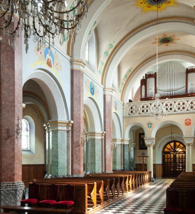 Zdjęcie nr 39: Budynki kościoła i plebanii znajdują się w centralnej części wsi, przy rynku na północ od głównej drogi z Krakowa do Oświęcimia. Kościół stoi przy obszernym placu, obwiedziony wysokim kamiennym murem z metalowym ogrodzeniem od południa. Kościół z prezbiterium zwróconym na północ, wzniesiony z kamienia wapiennego i cegły, tynkowany, z podmurówką. Świątynia wybudowana na planie krzyża łacińskiego z bazylikowym korpusem i transeptem. Dwuprzęsłowe prezbiterium zakończone półkoliście absydą zostało ujęte w dwa aneksy boczne, z których zachodni przeznaczony jest na zakrystię. Od południa do budynku przylega kruchta w przyziemiu wieży. Wnętrze kościoła trójnawowe, czteroprzęsłowe. Nawa główna łączy się z nawami bocznymi arkadami wspartymi na pięciobocznych filarach. Nawy boczne ujęte szerokimi pilastrami z pięciobocznymi filarami. Filary i pilastry stoją na wysokich, masywnych cokołach z kapitelami analogicznymi jak w nawie głównej. Ściany na skrzyżowaniu naw oraz ściany prezbiterium ujęte wspomnianymi filarami podtrzymującymi gurty sklepienia. Nawa i prezbiterium przykryte są sklepieniem kolebkowym z geometryzowanymi lunetami po obu stronach ścian. Ramiona transeptu sklepione kolebkowo z analogicznymi lunetami. Na skrzyżowaniu naw znajduje się sklepienie żaglaste. Nawę główną oświetlają rozmieszczone po jednym w każdym przęśle otwory okienne zamknięte łukiem półkolistym, okna w transepcie to biforia, widoczne tylko z zewnątrz okrągłe okno w absydzie wypełnione jest kamienną rozetą. Główne wejście od południa, do pierwszego przęsła od transeptu prowadzi dwoje drzwi w otworach o zamknięciu w kształcie łuku odciętego. W części zachodniej chór muzyczny wsparty na dwóch kolumnach i dwóch filarach dostawionych do ściany. Ich kapitele są analogiczne do pozostałych w kościele. Sklepienia dekorowane polichromią z rozetami i pękami lilii, a na skrzyżowaniu naw umieszczono malunek naścienny ukazujący Chrystusa tronującego z Bogiem Ojcem i gołębicą Ducha Świętego, a wokół nich cztery symbole ewangelistów. W fasadzie świątyni znajduje się strzelista wieża dwukondygnacyjna na planie czworokąta, przechodząca wyżej w ośmioboczną. Po bokach wieży wysokie aneksy zwieńczone schodkowo. Nad wejściem do kościoła znajduje się ozdobne okno zamknięte półkoliście i wypełnione czterema małymi rozetami ułożonymi jedna nad drugą. W kolejnej kondygnacji wieżę przepruwa biforium poniżej tarczy zegara i dekoracji arkadkowej. Wieża pełni jednocześnie funkcję dzwonnicy. Druga wieża na skrzyżowaniu nawy głównej i transeptu mieści sygnaturkę. Korpus kościoła nakryty dachem dwuspadowym, wieża wielobocznym. Wszystkie dachy kryte blachą.