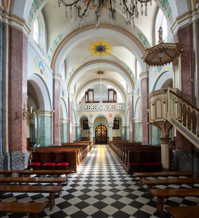 Zdjęcie nr 36: Budynki kościoła i plebanii znajdują się w centralnej części wsi, przy rynku na północ od głównej drogi z Krakowa do Oświęcimia. Kościół stoi przy obszernym placu, obwiedziony wysokim kamiennym murem z metalowym ogrodzeniem od południa. Kościół z prezbiterium zwróconym na północ, wzniesiony z kamienia wapiennego i cegły, tynkowany, z podmurówką. Świątynia wybudowana na planie krzyża łacińskiego z bazylikowym korpusem i transeptem. Dwuprzęsłowe prezbiterium zakończone półkoliście absydą zostało ujęte w dwa aneksy boczne, z których zachodni przeznaczony jest na zakrystię. Od południa do budynku przylega kruchta w przyziemiu wieży. Wnętrze kościoła trójnawowe, czteroprzęsłowe. Nawa główna łączy się z nawami bocznymi arkadami wspartymi na pięciobocznych filarach. Nawy boczne ujęte szerokimi pilastrami z pięciobocznymi filarami. Filary i pilastry stoją na wysokich, masywnych cokołach z kapitelami analogicznymi jak w nawie głównej. Ściany na skrzyżowaniu naw oraz ściany prezbiterium ujęte wspomnianymi filarami podtrzymującymi gurty sklepienia. Nawa i prezbiterium przykryte są sklepieniem kolebkowym z geometryzowanymi lunetami po obu stronach ścian. Ramiona transeptu sklepione kolebkowo z analogicznymi lunetami. Na skrzyżowaniu naw znajduje się sklepienie żaglaste. Nawę główną oświetlają rozmieszczone po jednym w każdym przęśle otwory okienne zamknięte łukiem półkolistym, okna w transepcie to biforia, widoczne tylko z zewnątrz okrągłe okno w absydzie wypełnione jest kamienną rozetą. Główne wejście od południa, do pierwszego przęsła od transeptu prowadzi dwoje drzwi w otworach o zamknięciu w kształcie łuku odciętego. W części zachodniej chór muzyczny wsparty na dwóch kolumnach i dwóch filarach dostawionych do ściany. Ich kapitele są analogiczne do pozostałych w kościele. Sklepienia dekorowane polichromią z rozetami i pękami lilii, a na skrzyżowaniu naw umieszczono malunek naścienny ukazujący Chrystusa tronującego z Bogiem Ojcem i gołębicą Ducha Świętego, a wokół nich cztery symbole ewangelistów. W fasadzie świątyni znajduje się strzelista wieża dwukondygnacyjna na planie czworokąta, przechodząca wyżej w ośmioboczną. Po bokach wieży wysokie aneksy zwieńczone schodkowo. Nad wejściem do kościoła znajduje się ozdobne okno zamknięte półkoliście i wypełnione czterema małymi rozetami ułożonymi jedna nad drugą. W kolejnej kondygnacji wieżę przepruwa biforium poniżej tarczy zegara i dekoracji arkadkowej. Wieża pełni jednocześnie funkcję dzwonnicy. Druga wieża na skrzyżowaniu nawy głównej i transeptu mieści sygnaturkę. Korpus kościoła nakryty dachem dwuspadowym, wieża wielobocznym. Wszystkie dachy kryte blachą.