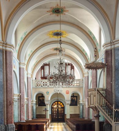 Zdjęcie nr 37: Budynki kościoła i plebanii znajdują się w centralnej części wsi, przy rynku na północ od głównej drogi z Krakowa do Oświęcimia. Kościół stoi przy obszernym placu, obwiedziony wysokim kamiennym murem z metalowym ogrodzeniem od południa. Kościół z prezbiterium zwróconym na północ, wzniesiony z kamienia wapiennego i cegły, tynkowany, z podmurówką. Świątynia wybudowana na planie krzyża łacińskiego z bazylikowym korpusem i transeptem. Dwuprzęsłowe prezbiterium zakończone półkoliście absydą zostało ujęte w dwa aneksy boczne, z których zachodni przeznaczony jest na zakrystię. Od południa do budynku przylega kruchta w przyziemiu wieży. Wnętrze kościoła trójnawowe, czteroprzęsłowe. Nawa główna łączy się z nawami bocznymi arkadami wspartymi na pięciobocznych filarach. Nawy boczne ujęte szerokimi pilastrami z pięciobocznymi filarami. Filary i pilastry stoją na wysokich, masywnych cokołach z kapitelami analogicznymi jak w nawie głównej. Ściany na skrzyżowaniu naw oraz ściany prezbiterium ujęte wspomnianymi filarami podtrzymującymi gurty sklepienia. Nawa i prezbiterium przykryte są sklepieniem kolebkowym z geometryzowanymi lunetami po obu stronach ścian. Ramiona transeptu sklepione kolebkowo z analogicznymi lunetami. Na skrzyżowaniu naw znajduje się sklepienie żaglaste. Nawę główną oświetlają rozmieszczone po jednym w każdym przęśle otwory okienne zamknięte łukiem półkolistym, okna w transepcie to biforia, widoczne tylko z zewnątrz okrągłe okno w absydzie wypełnione jest kamienną rozetą. Główne wejście od południa, do pierwszego przęsła od transeptu prowadzi dwoje drzwi w otworach o zamknięciu w kształcie łuku odciętego. W części zachodniej chór muzyczny wsparty na dwóch kolumnach i dwóch filarach dostawionych do ściany. Ich kapitele są analogiczne do pozostałych w kościele. Sklepienia dekorowane polichromią z rozetami i pękami lilii, a na skrzyżowaniu naw umieszczono malunek naścienny ukazujący Chrystusa tronującego z Bogiem Ojcem i gołębicą Ducha Świętego, a wokół nich cztery symbole ewangelistów. W fasadzie świątyni znajduje się strzelista wieża dwukondygnacyjna na planie czworokąta, przechodząca wyżej w ośmioboczną. Po bokach wieży wysokie aneksy zwieńczone schodkowo. Nad wejściem do kościoła znajduje się ozdobne okno zamknięte półkoliście i wypełnione czterema małymi rozetami ułożonymi jedna nad drugą. W kolejnej kondygnacji wieżę przepruwa biforium poniżej tarczy zegara i dekoracji arkadkowej. Wieża pełni jednocześnie funkcję dzwonnicy. Druga wieża na skrzyżowaniu nawy głównej i transeptu mieści sygnaturkę. Korpus kościoła nakryty dachem dwuspadowym, wieża wielobocznym. Wszystkie dachy kryte blachą.