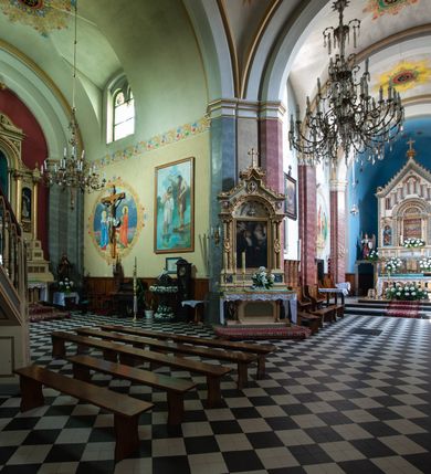 Zdjęcie nr 32: Budynki kościoła i plebanii znajdują się w centralnej części wsi, przy rynku na północ od głównej drogi z Krakowa do Oświęcimia. Kościół stoi przy obszernym placu, obwiedziony wysokim kamiennym murem z metalowym ogrodzeniem od południa. Kościół z prezbiterium zwróconym na północ, wzniesiony z kamienia wapiennego i cegły, tynkowany, z podmurówką. Świątynia wybudowana na planie krzyża łacińskiego z bazylikowym korpusem i transeptem. Dwuprzęsłowe prezbiterium zakończone półkoliście absydą zostało ujęte w dwa aneksy boczne, z których zachodni przeznaczony jest na zakrystię. Od południa do budynku przylega kruchta w przyziemiu wieży. Wnętrze kościoła trójnawowe, czteroprzęsłowe. Nawa główna łączy się z nawami bocznymi arkadami wspartymi na pięciobocznych filarach. Nawy boczne ujęte szerokimi pilastrami z pięciobocznymi filarami. Filary i pilastry stoją na wysokich, masywnych cokołach z kapitelami analogicznymi jak w nawie głównej. Ściany na skrzyżowaniu naw oraz ściany prezbiterium ujęte wspomnianymi filarami podtrzymującymi gurty sklepienia. Nawa i prezbiterium przykryte są sklepieniem kolebkowym z geometryzowanymi lunetami po obu stronach ścian. Ramiona transeptu sklepione kolebkowo z analogicznymi lunetami. Na skrzyżowaniu naw znajduje się sklepienie żaglaste. Nawę główną oświetlają rozmieszczone po jednym w każdym przęśle otwory okienne zamknięte łukiem półkolistym, okna w transepcie to biforia, widoczne tylko z zewnątrz okrągłe okno w absydzie wypełnione jest kamienną rozetą. Główne wejście od południa, do pierwszego przęsła od transeptu prowadzi dwoje drzwi w otworach o zamknięciu w kształcie łuku odciętego. W części zachodniej chór muzyczny wsparty na dwóch kolumnach i dwóch filarach dostawionych do ściany. Ich kapitele są analogiczne do pozostałych w kościele. Sklepienia dekorowane polichromią z rozetami i pękami lilii, a na skrzyżowaniu naw umieszczono malunek naścienny ukazujący Chrystusa tronującego z Bogiem Ojcem i gołębicą Ducha Świętego, a wokół nich cztery symbole ewangelistów. W fasadzie świątyni znajduje się strzelista wieża dwukondygnacyjna na planie czworokąta, przechodząca wyżej w ośmioboczną. Po bokach wieży wysokie aneksy zwieńczone schodkowo. Nad wejściem do kościoła znajduje się ozdobne okno zamknięte półkoliście i wypełnione czterema małymi rozetami ułożonymi jedna nad drugą. W kolejnej kondygnacji wieżę przepruwa biforium poniżej tarczy zegara i dekoracji arkadkowej. Wieża pełni jednocześnie funkcję dzwonnicy. Druga wieża na skrzyżowaniu nawy głównej i transeptu mieści sygnaturkę. Korpus kościoła nakryty dachem dwuspadowym, wieża wielobocznym. Wszystkie dachy kryte blachą.