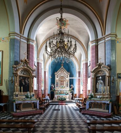 Zdjęcie nr 30: Budynki kościoła i plebanii znajdują się w centralnej części wsi, przy rynku na północ od głównej drogi z Krakowa do Oświęcimia. Kościół stoi przy obszernym placu, obwiedziony wysokim kamiennym murem z metalowym ogrodzeniem od południa. Kościół z prezbiterium zwróconym na północ, wzniesiony z kamienia wapiennego i cegły, tynkowany, z podmurówką. Świątynia wybudowana na planie krzyża łacińskiego z bazylikowym korpusem i transeptem. Dwuprzęsłowe prezbiterium zakończone półkoliście absydą zostało ujęte w dwa aneksy boczne, z których zachodni przeznaczony jest na zakrystię. Od południa do budynku przylega kruchta w przyziemiu wieży. Wnętrze kościoła trójnawowe, czteroprzęsłowe. Nawa główna łączy się z nawami bocznymi arkadami wspartymi na pięciobocznych filarach. Nawy boczne ujęte szerokimi pilastrami z pięciobocznymi filarami. Filary i pilastry stoją na wysokich, masywnych cokołach z kapitelami analogicznymi jak w nawie głównej. Ściany na skrzyżowaniu naw oraz ściany prezbiterium ujęte wspomnianymi filarami podtrzymującymi gurty sklepienia. Nawa i prezbiterium przykryte są sklepieniem kolebkowym z geometryzowanymi lunetami po obu stronach ścian. Ramiona transeptu sklepione kolebkowo z analogicznymi lunetami. Na skrzyżowaniu naw znajduje się sklepienie żaglaste. Nawę główną oświetlają rozmieszczone po jednym w każdym przęśle otwory okienne zamknięte łukiem półkolistym, okna w transepcie to biforia, widoczne tylko z zewnątrz okrągłe okno w absydzie wypełnione jest kamienną rozetą. Główne wejście od południa, do pierwszego przęsła od transeptu prowadzi dwoje drzwi w otworach o zamknięciu w kształcie łuku odciętego. W części zachodniej chór muzyczny wsparty na dwóch kolumnach i dwóch filarach dostawionych do ściany. Ich kapitele są analogiczne do pozostałych w kościele. Sklepienia dekorowane polichromią z rozetami i pękami lilii, a na skrzyżowaniu naw umieszczono malunek naścienny ukazujący Chrystusa tronującego z Bogiem Ojcem i gołębicą Ducha Świętego, a wokół nich cztery symbole ewangelistów. W fasadzie świątyni znajduje się strzelista wieża dwukondygnacyjna na planie czworokąta, przechodząca wyżej w ośmioboczną. Po bokach wieży wysokie aneksy zwieńczone schodkowo. Nad wejściem do kościoła znajduje się ozdobne okno zamknięte półkoliście i wypełnione czterema małymi rozetami ułożonymi jedna nad drugą. W kolejnej kondygnacji wieżę przepruwa biforium poniżej tarczy zegara i dekoracji arkadkowej. Wieża pełni jednocześnie funkcję dzwonnicy. Druga wieża na skrzyżowaniu nawy głównej i transeptu mieści sygnaturkę. Korpus kościoła nakryty dachem dwuspadowym, wieża wielobocznym. Wszystkie dachy kryte blachą.