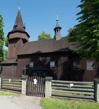 Zdjęcie nr 7: Kościół św. Jakuba w Palczowicach został wybudowany w roku 1894 w miejsce wcześniejszego kościoła z roku 1498. Kościół halowy, na kamiennej podmurówce, wzniesiony z drewna.  Kościół jest jednonawowy, orientowany.  Po jego zachodniej stronie szeroka, czworoboczna wieża nakryta ośmiobocznym hełmem. Pod hełmem również ośmioboczny szeroki, drewniany kandelabr. Hełm strzelisty, kryty gontem, zwieńczony metalowym krzyżem. Z czterech stron hełmu metalowe wykusze okienne  ze stylizowanymi sterczynami. Po czterech stronach korpusu wieży w jego wysokości małe otwory okienne  zwieńczone ostrokątnie. Korpus halowy nakryty dwuspadowym dachem z gontu.  We wschodniej części dachu ośmioboczna metalowa sygnaturka również zwieńczona krzyżem. Jej hełm strzelisty z pokryciem metalowym. W korpusie od południa i północy po dwa prostokątne otwory okienne z bliźniaczymi zamkniętymi ostrołukowo kwaterami. Prezbiterium niższe i węższe w stosunku do nawy, kryte osobnym, dwuspadowym dachem gontowym, zamknięte trójbocznie. Po południowej stronie prezbiterium dwa otwory okienne analogiczne do zlokalizowanych w nawie. Po północnej stronie prezbiterium dobudowana murowana, tynkowana zakrystia z dachem jednospadowym, krytym blachą.  W zakrystii jeden czworokątny otwór okienny i osobne wejście po stronie wschodniej. Nad głównym wejściem do kościoła, z czworobocznymi, dwuskrzydłowymi drzwiami  wysunięte dwuspadowe zadaszenie Po bokach drzwi w połowie ich wysokości symetrycznie ułożone, czworokątne otwory okienne. Do południowej ściany nawy głównej przylegająca drewniana kruchta na planie prostokątnym z dwuspadowym zadaszeniem zwieńczonym krzyżem. Po obu stronach drzwi otwory okienne zwieńczone ostrokątnie.