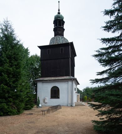 Zdjęcie nr 3: Dzwonnica na planie kwadratu do połowy wysokości murowana, wyżej drewniana, zwieńczona baniastym hełmem z latarnią. 
Część murowana nieco szersza. Od strony wschodniej znajdują się drzwi wejściowe, naroża są wzmocnione lizenami, od północy i południa znajdują się okna prostokątne zamknięte łukiem półokrągłym. Kondygnacje oddziela szeroki fartuch. Górna partia z pozorną izbicą przeprutą z każdej strony dwoma niewielkimi oknami w kształcie stojącego prostokąta zamkniętego łukiem pełnym. Od strony południowej otwór drzwiowy prostokątny zamknięty łukiem półokrągłym. Hełm baniasty przechodzi w oktogon,  na którym są arkadki wspierające mniejszy hełm cebulasty zwieńczony krzyżem. Dachy kryte blachą. 
